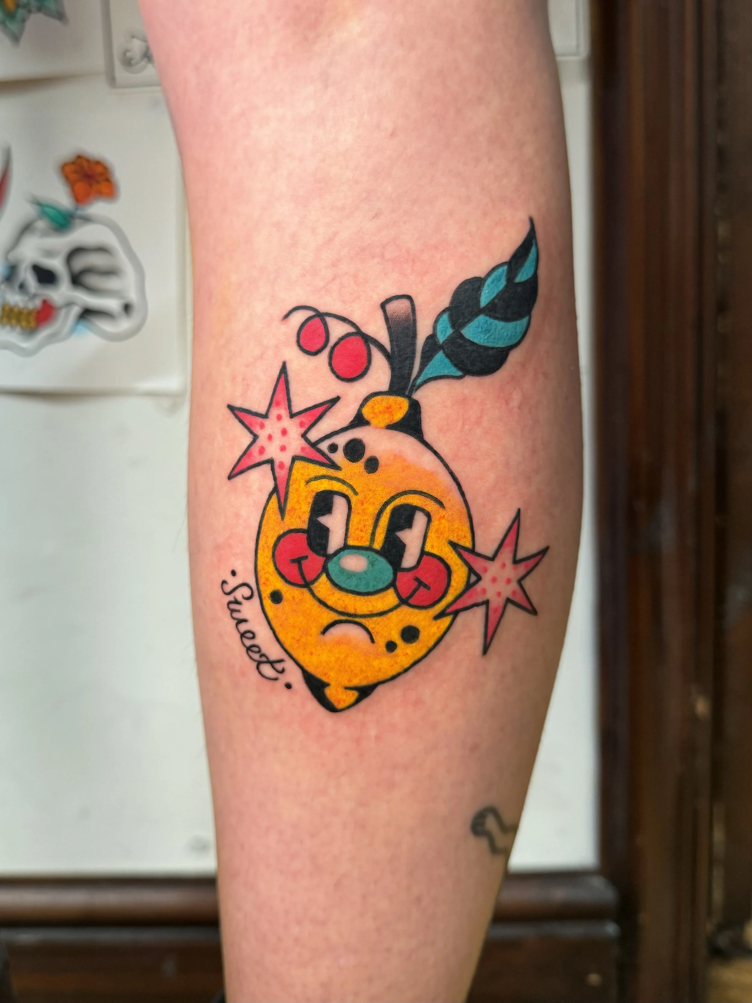 Colorful tattoo of a cartoon-style  Lemon with a face with a mischievous expression, surrounded by stars, cherries, and leaves, with the word 'Sweeet' written in cursive.