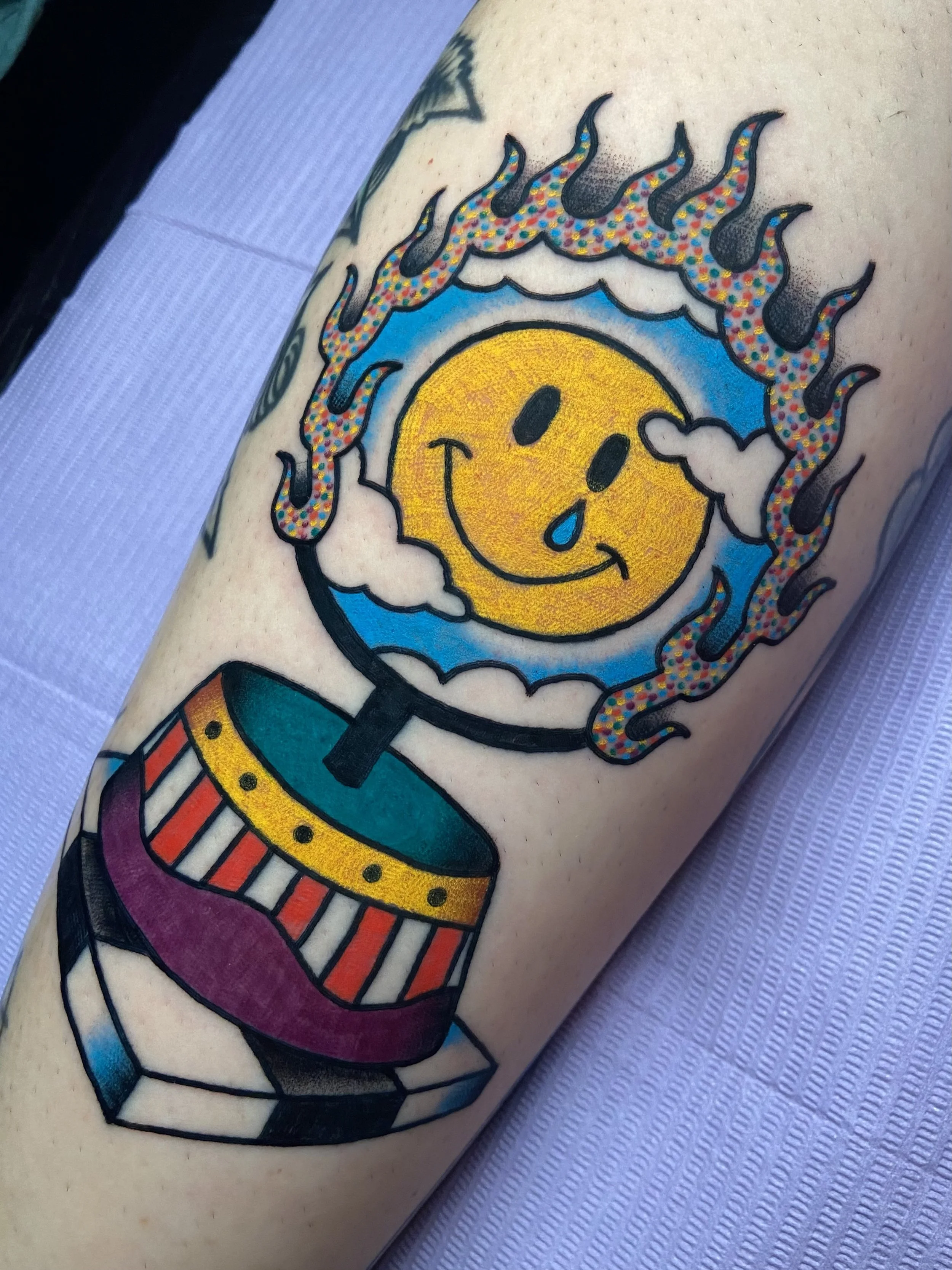 Tattoo of a smiling yellow sun with a tear and a happy face, in a flaming circus hoop with a striped pattern, surrounded by multicolored flames.