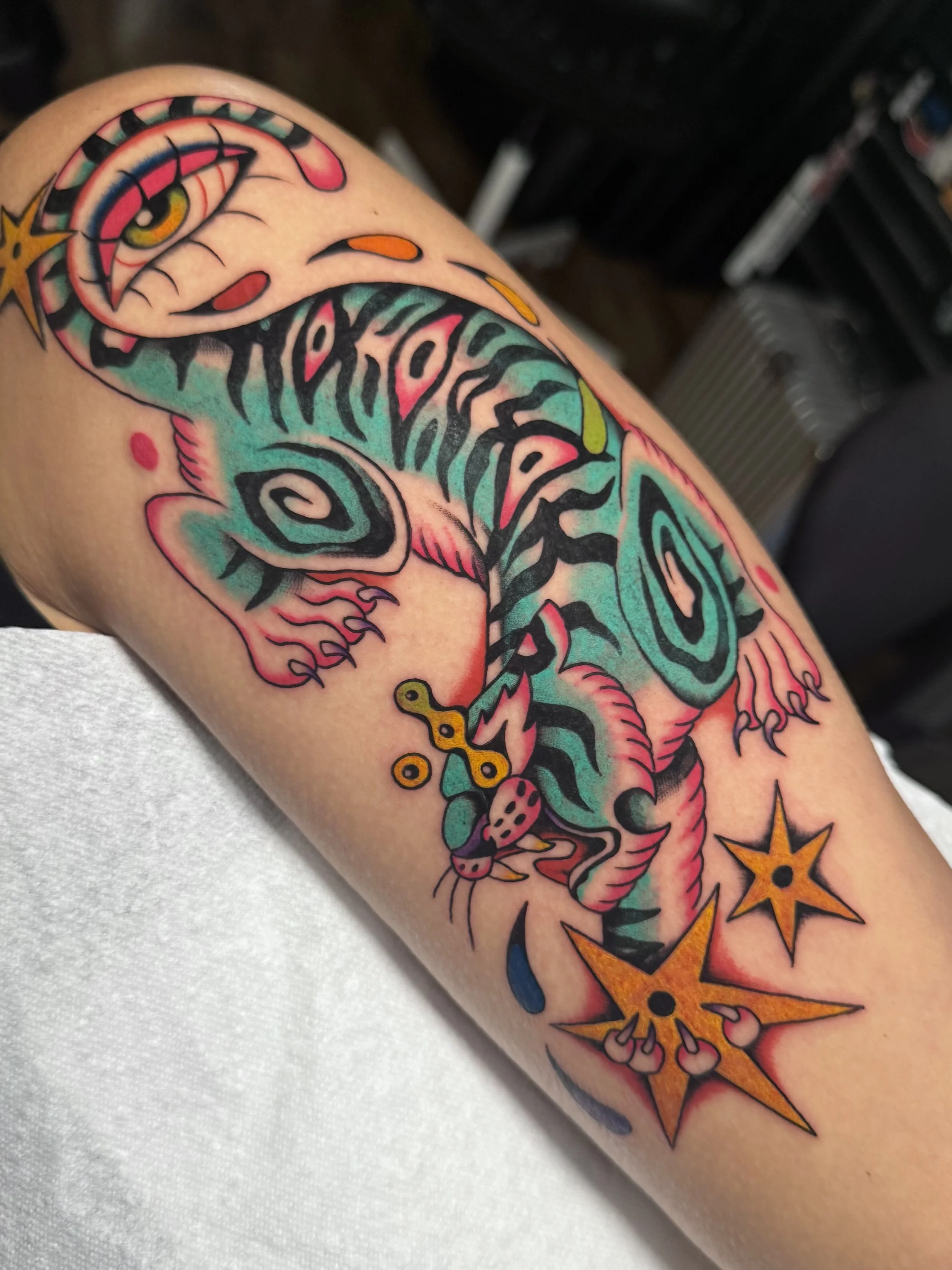 Colorful tattoo of a tiger with pink, turquoise, and black stripes, surrounded by yellow stars and abstract shapes on a person's upper arm.