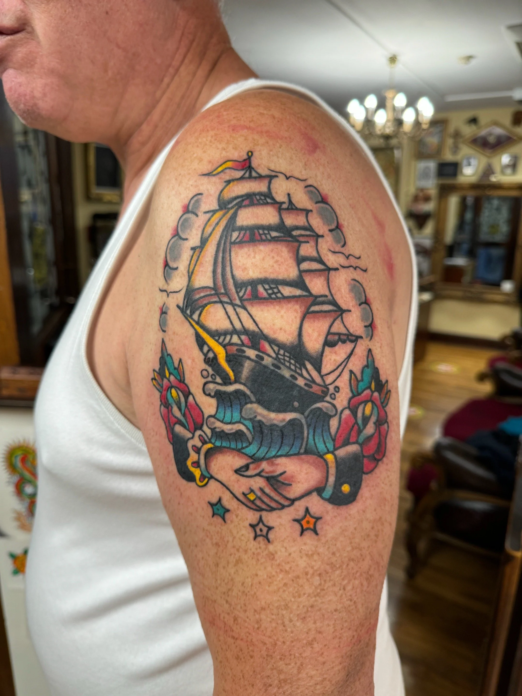 A tattoo on a person's upper arm depicting a traditional pirate ship sailing on water, with clouds and a flag at the top, surrounded by colorful roses, a handshake, and star shapes.