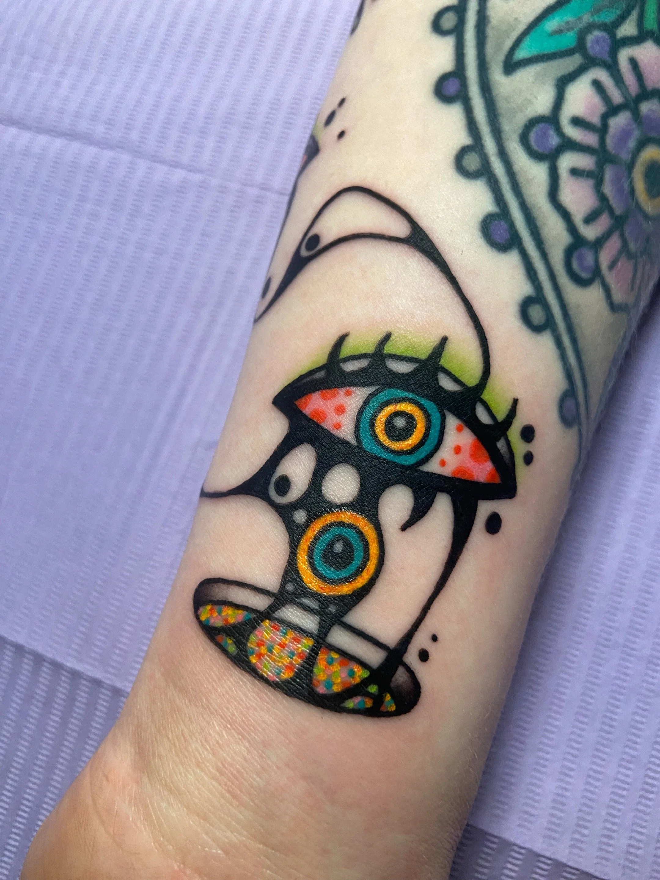 A colorful tattoo featuring an eye with vibrant patterns inside, connected by a symmetrical black design with colorful circular elements at both ends, on a person's arm.