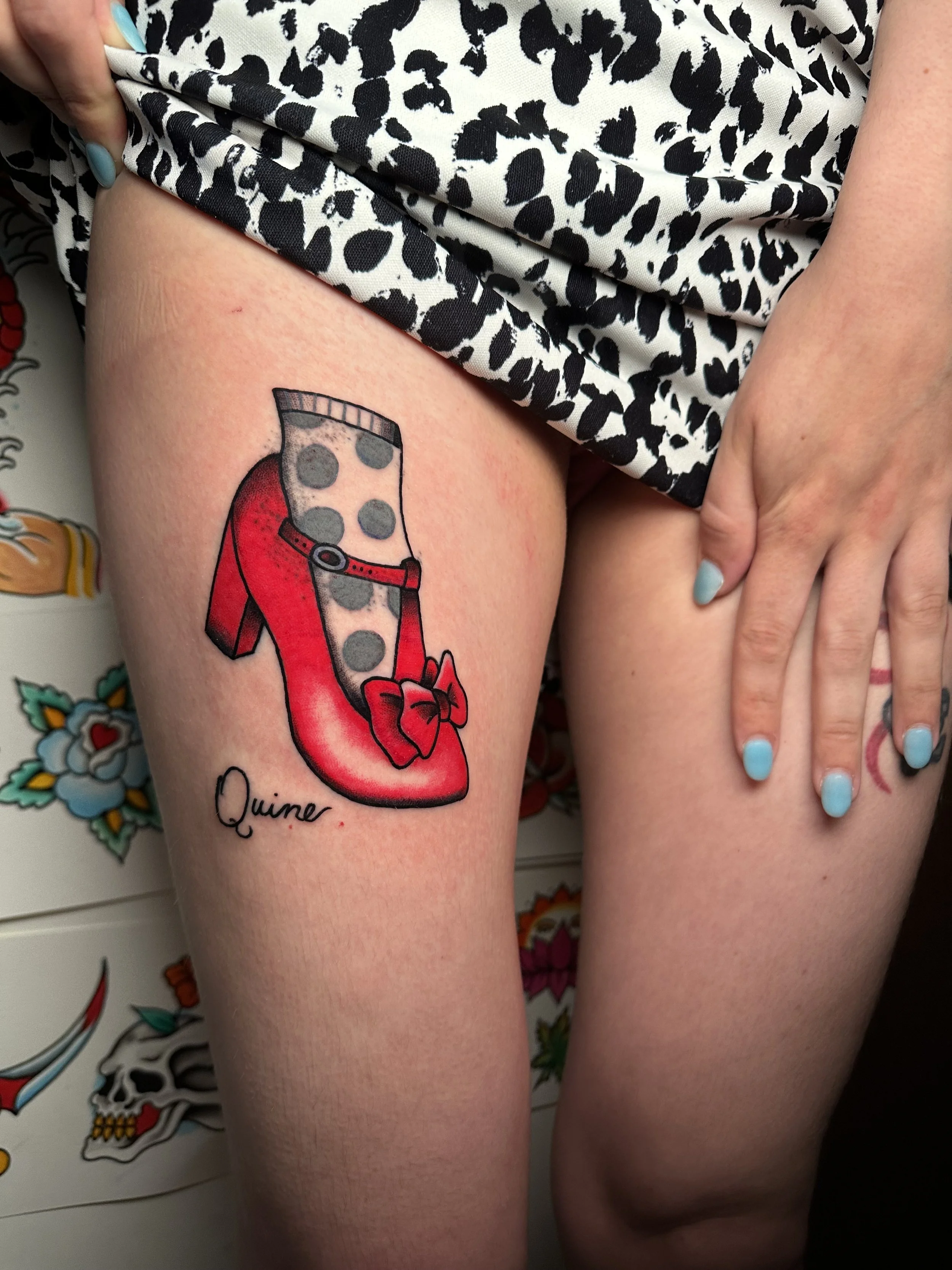 Tattoo of a high-heeled shoe with a polka dot stocking and a red bow, labeled "Quine," on a person's thigh.