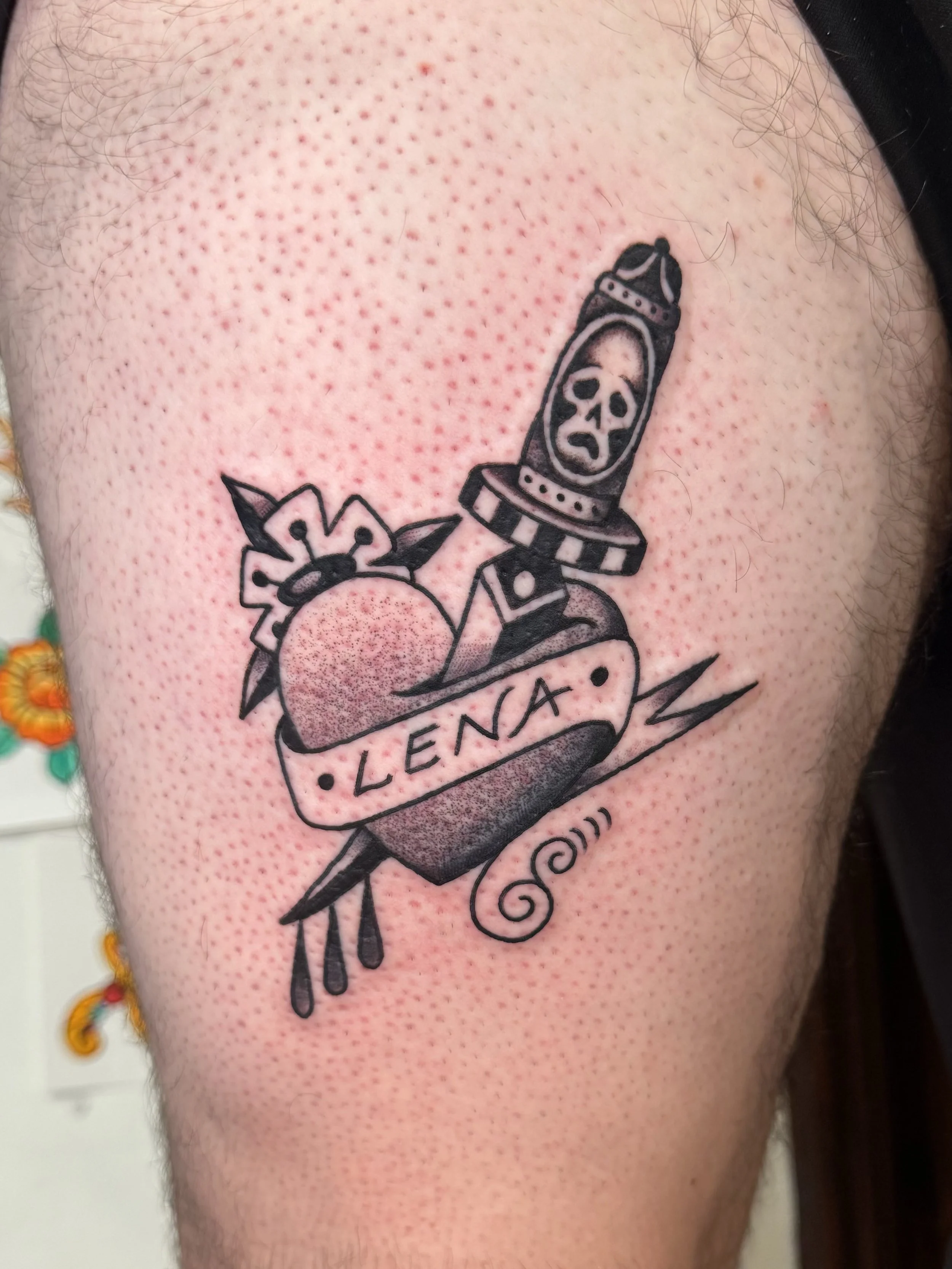 A traditional tattoo on a person's thigh depicts a heart with a banner reading 'LENA', topped with a rose, a dagger with a skull inside, and decorative leaves and swirls.