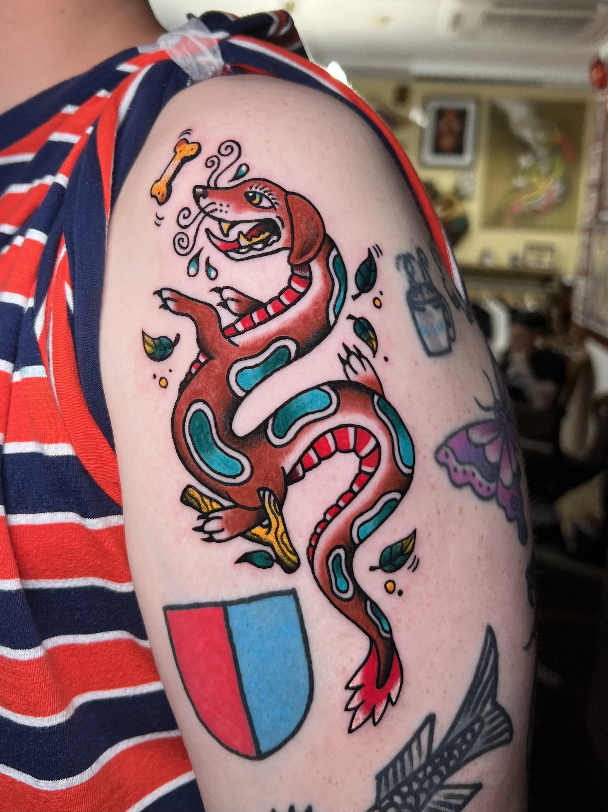 Tattoo of a cartoon-style dragon dog with a smiling face, wings, and a forked tongue, wrapped around a shield divided into red and blue sections.