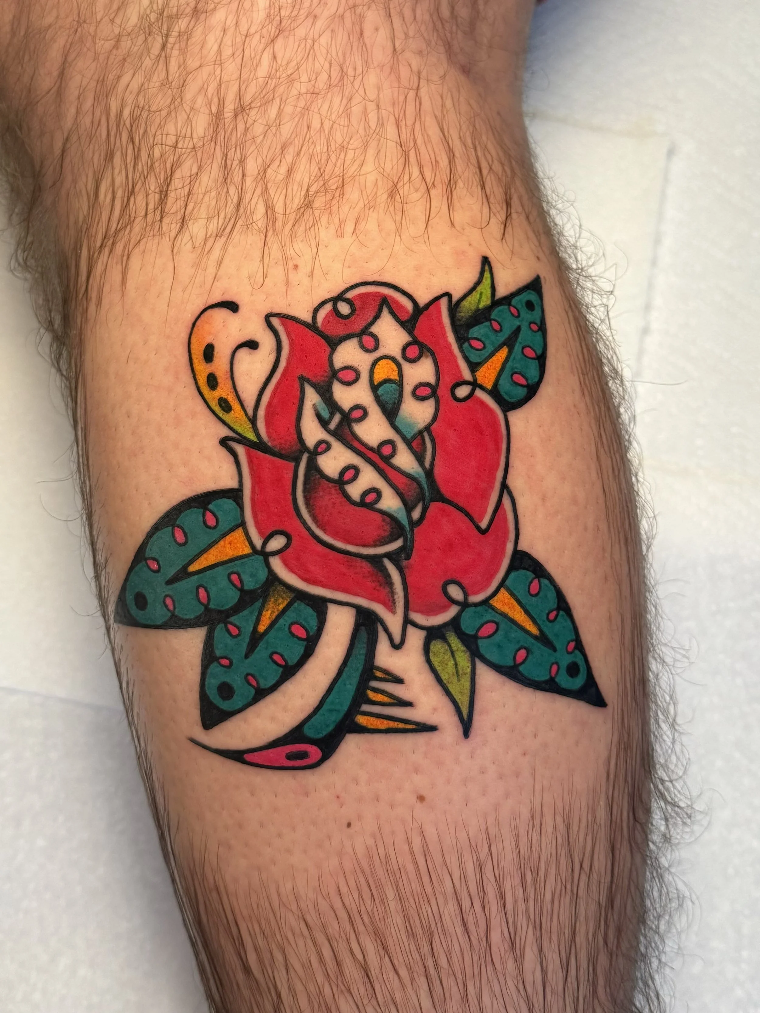 Colorful tattoo of a rose with leaves, the tattoo is traditional inspired, but has extra accents and various decorative elements on skin.