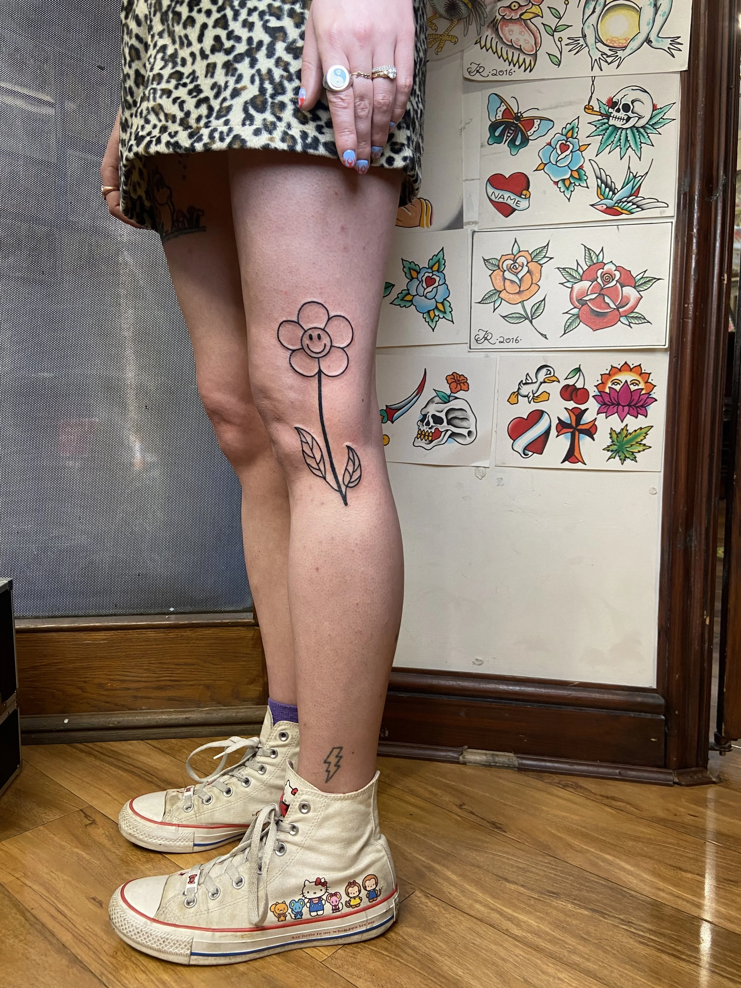 A woman wearing a leopard print dress, showing her tattoos on her leg and ankle. The tattoo on her thigh is a simple black outline of a flower with a smiling face. The tattoo on her ankle is a black lightning bolt. She is standing on a wooden floor, 