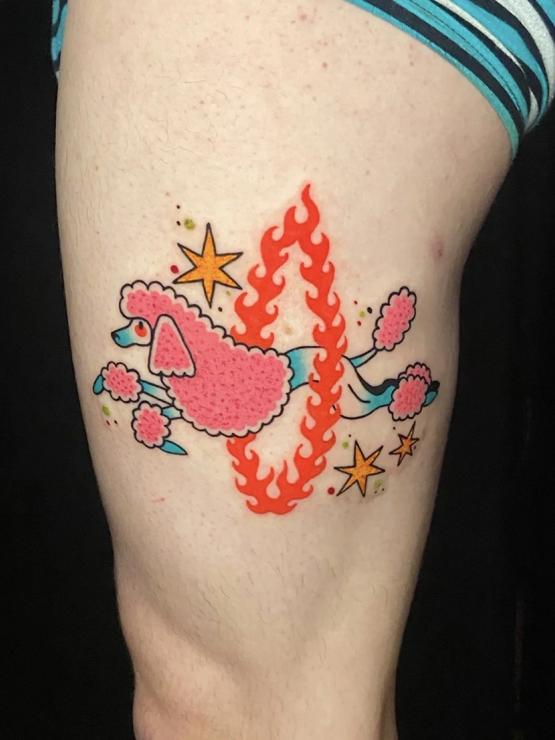 Tattoo of a pink poodle with blue ears and tail, surrounded by orange flames, yellow stars, pink clouds, and small green and pink dots on person's thigh.