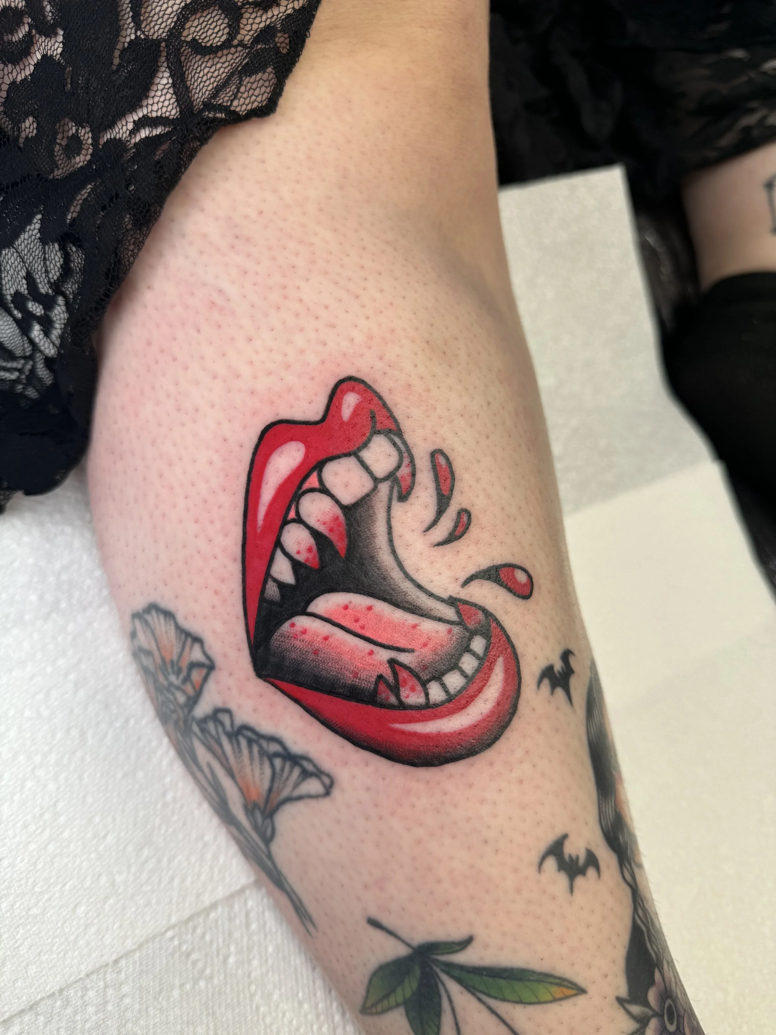 Tattoo of an open vampire mouth with red lips, black and gray interior, and sharp teeth on a person's upper arm.