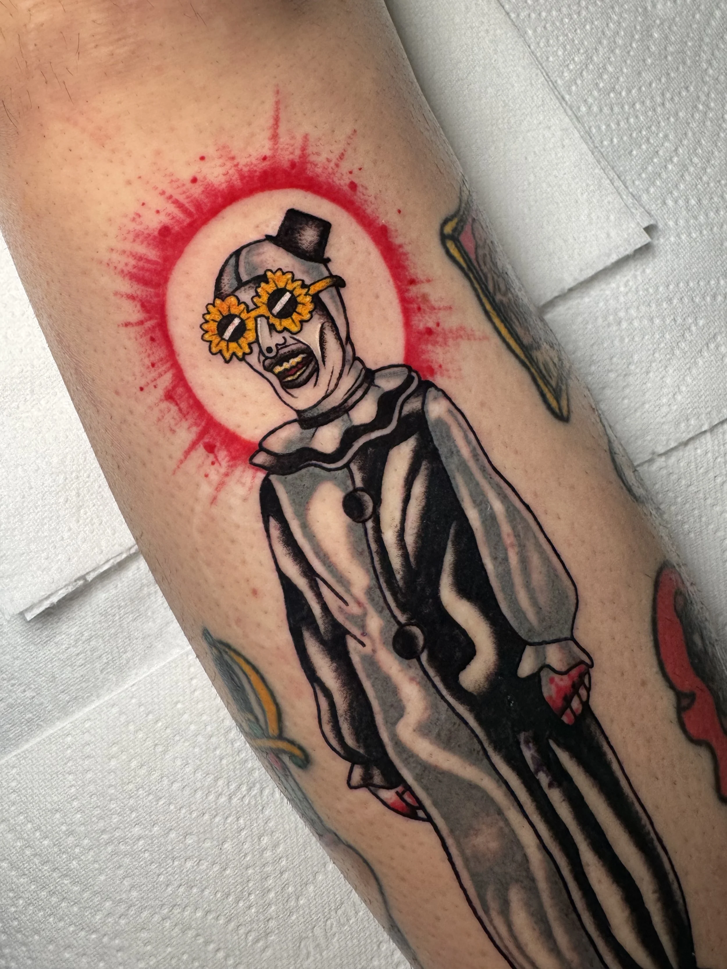 Tattoo of art the clown from the horror film the terrifier with sunflower sunglasses, a top hat, and a striped outfit, with a red  blood spatter halo behind its head.