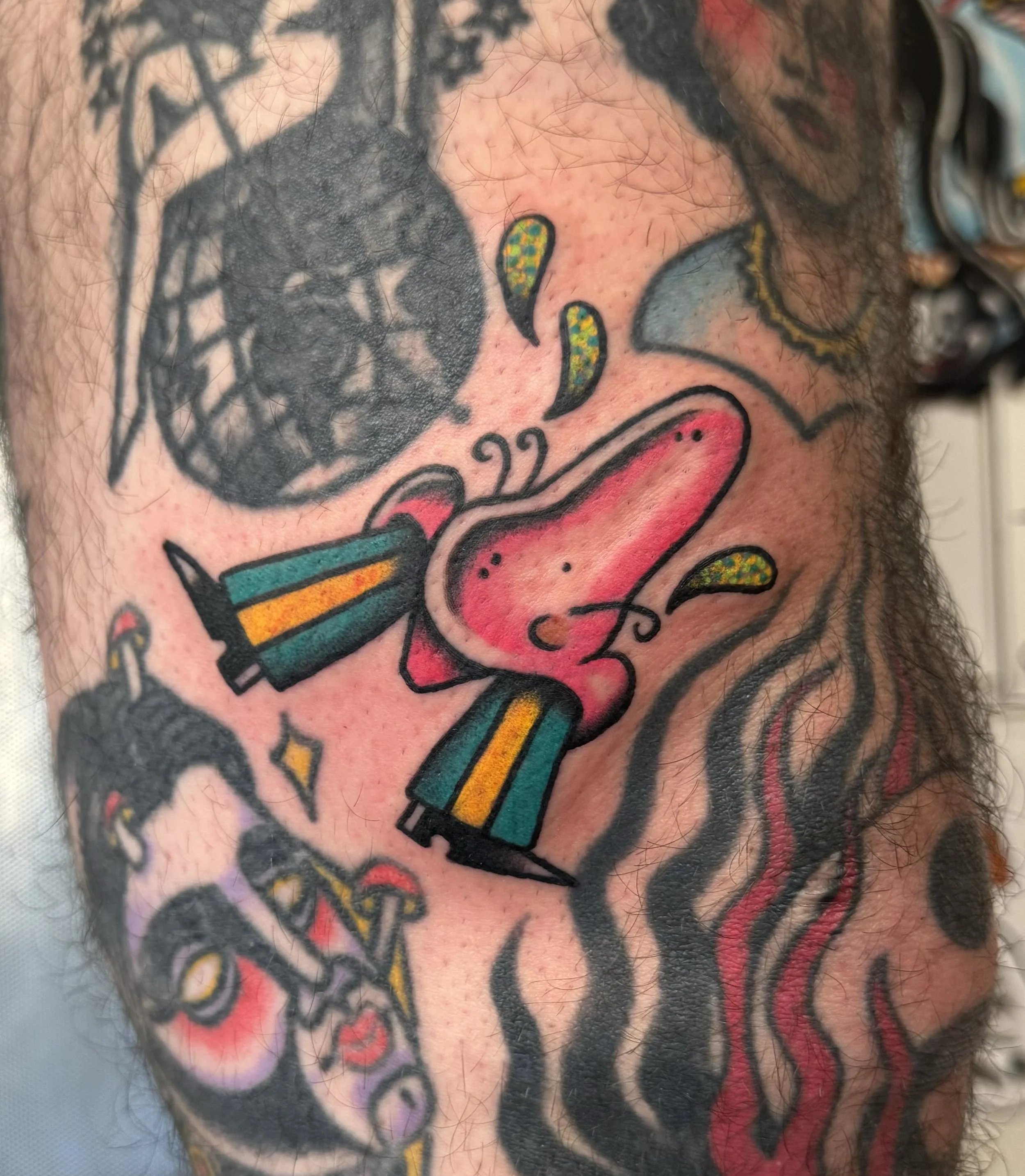 Colorful tattoo of a nose wearing trousers, with red and black accents, surrounded by black and green abstract elements and flames.