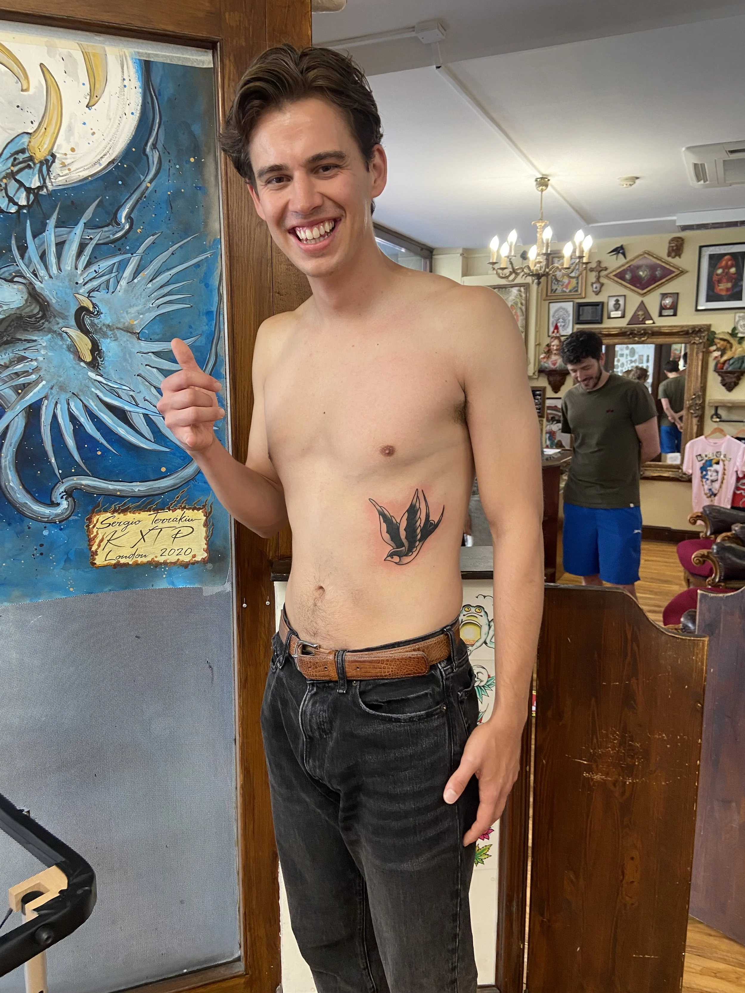 A shirtless young man with dark hair and a smile, pointing at a tattoo of a swallow on his side, standing inside a tattoo parlour.