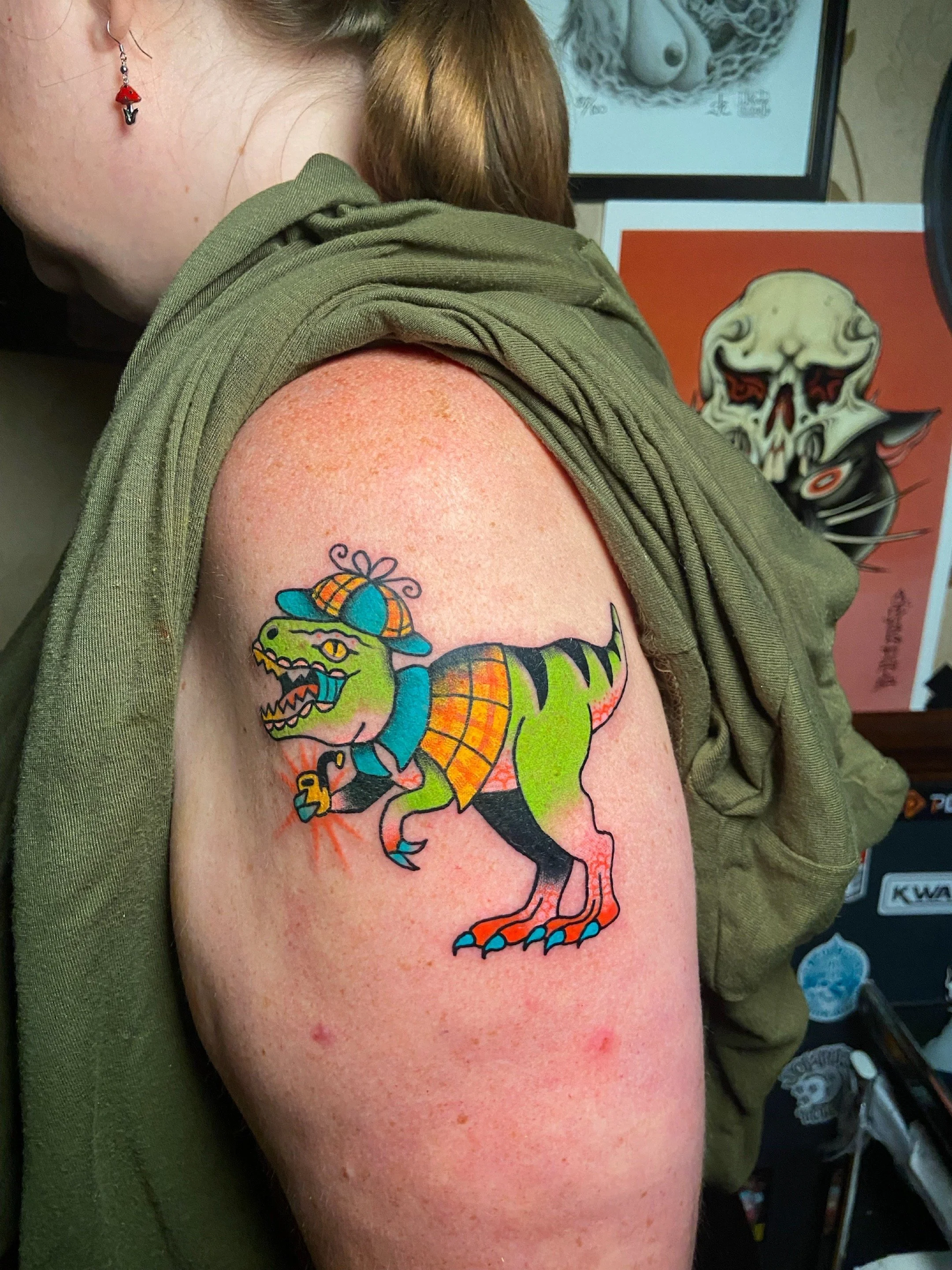 Colorful tattoo of a dinosaur wearing a hat and tie on person's upper arm.