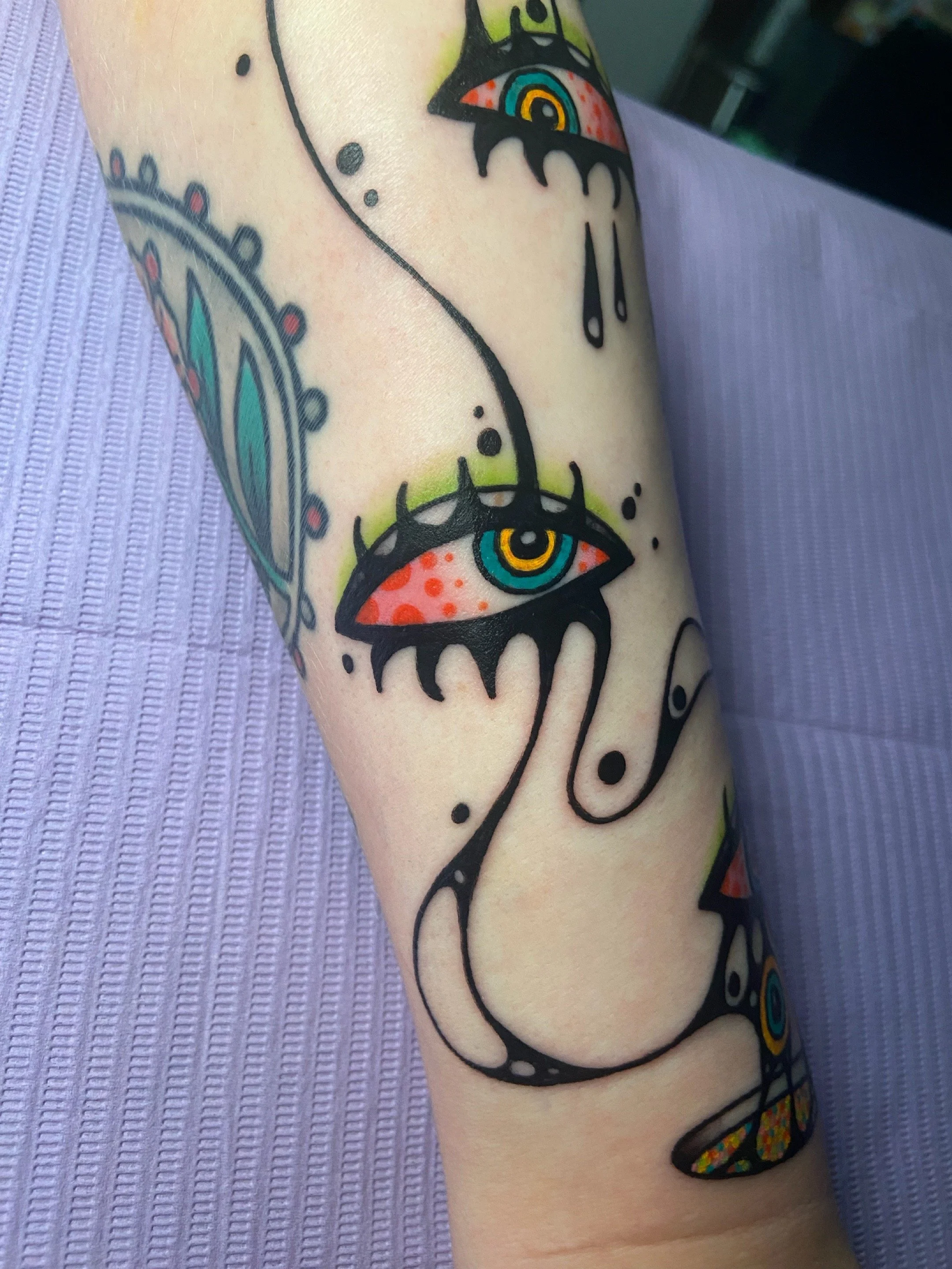 Tattoo of stylized, colorful eyes with black lashes, dripping ink, and abstract lines on a person's forearm.