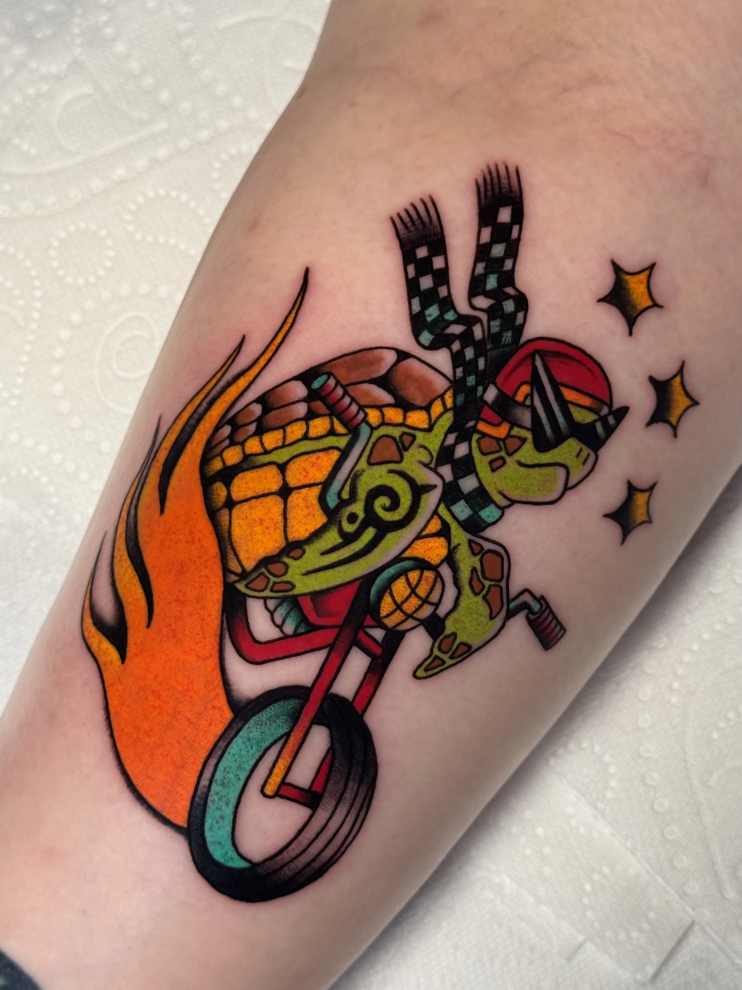 Tattoo of a cartoon-style turtle riding a red motorcycle with flames and stars around it.