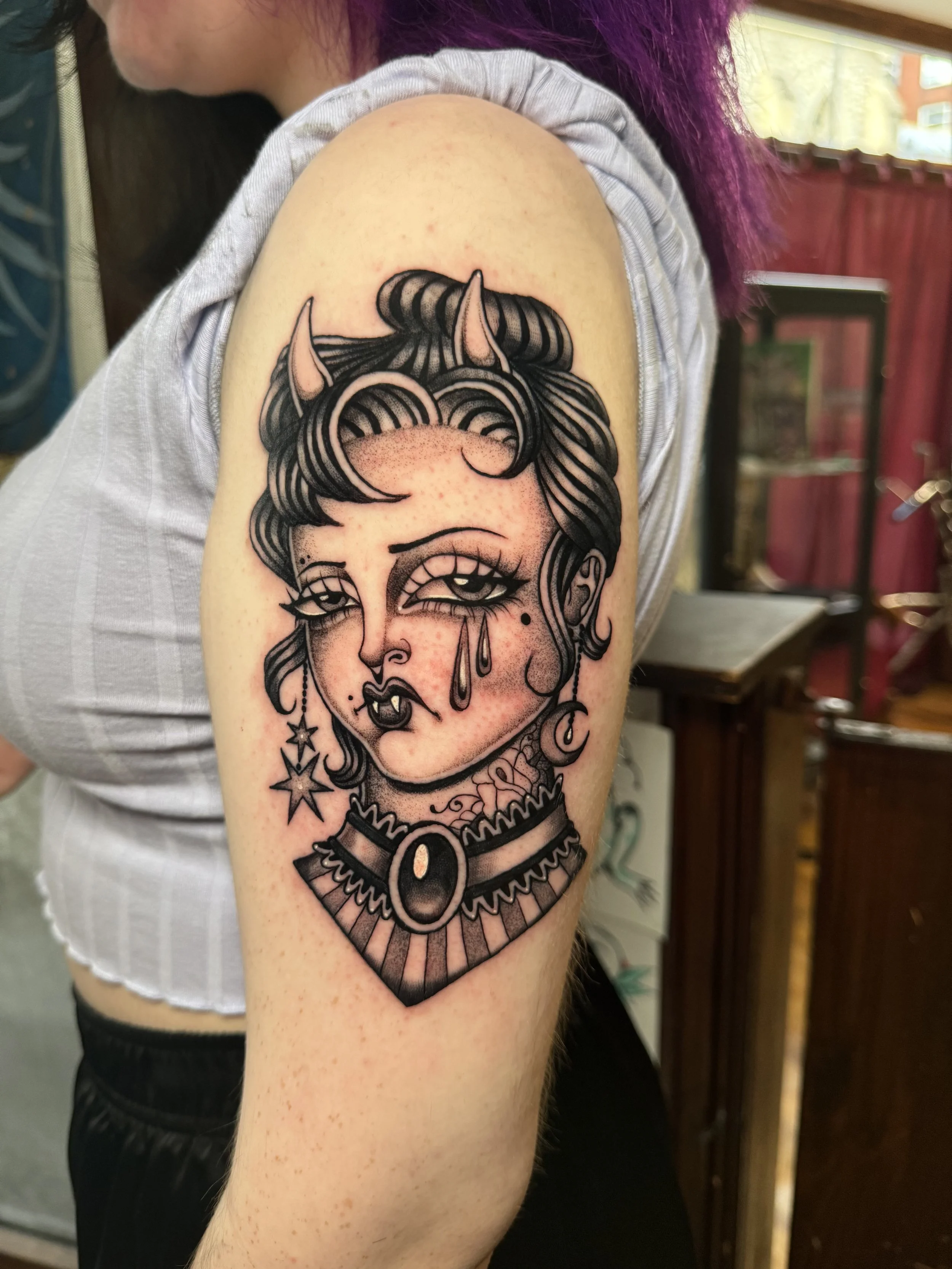 Tattoo of a stylized woman's face with horns, teardrops, and gothic jewelry on upper arm.