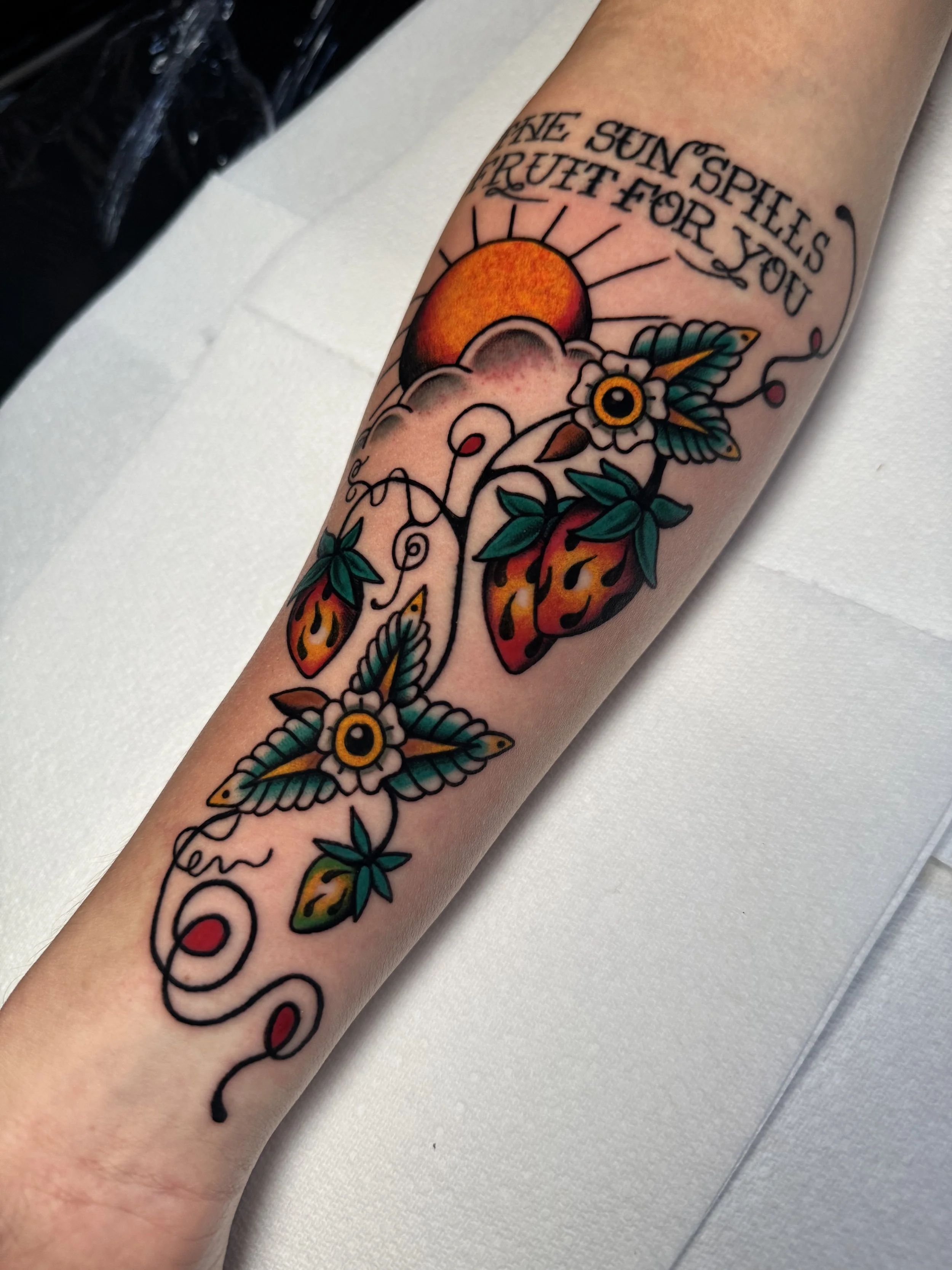 Colorful tattoo on a person's forearm featuring a sun, clouds, flowers, and butterflies, with the text "The sun spills fruit for you" at the top.