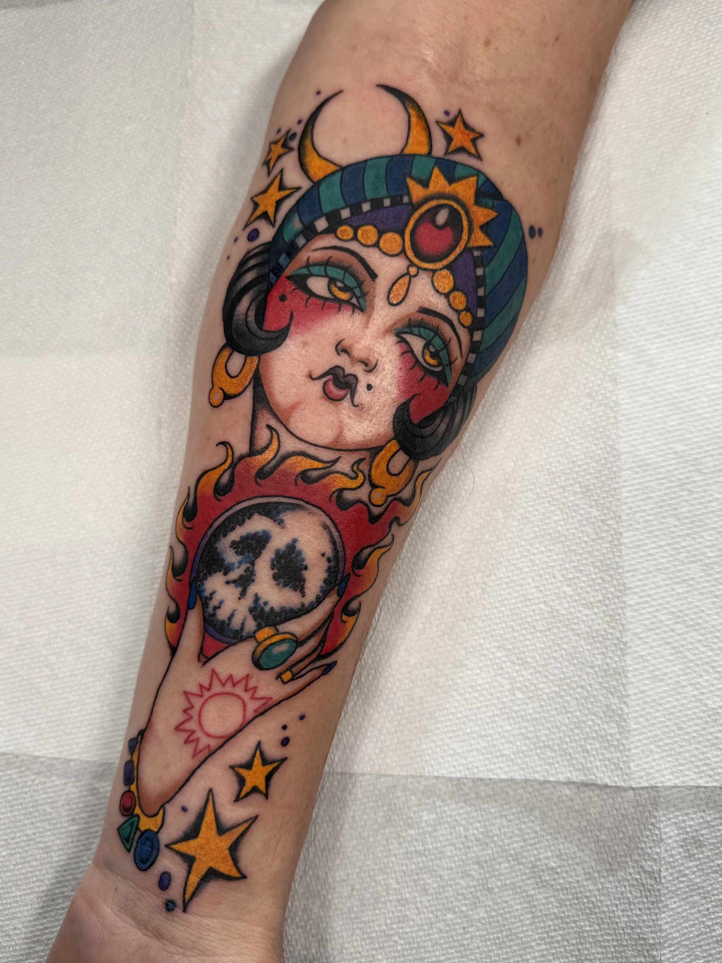 Colorful tattoo of a mystical gypsy woman with a moon, holding a crystal ball, surrounded by stars, flames, and cosmic symbols.