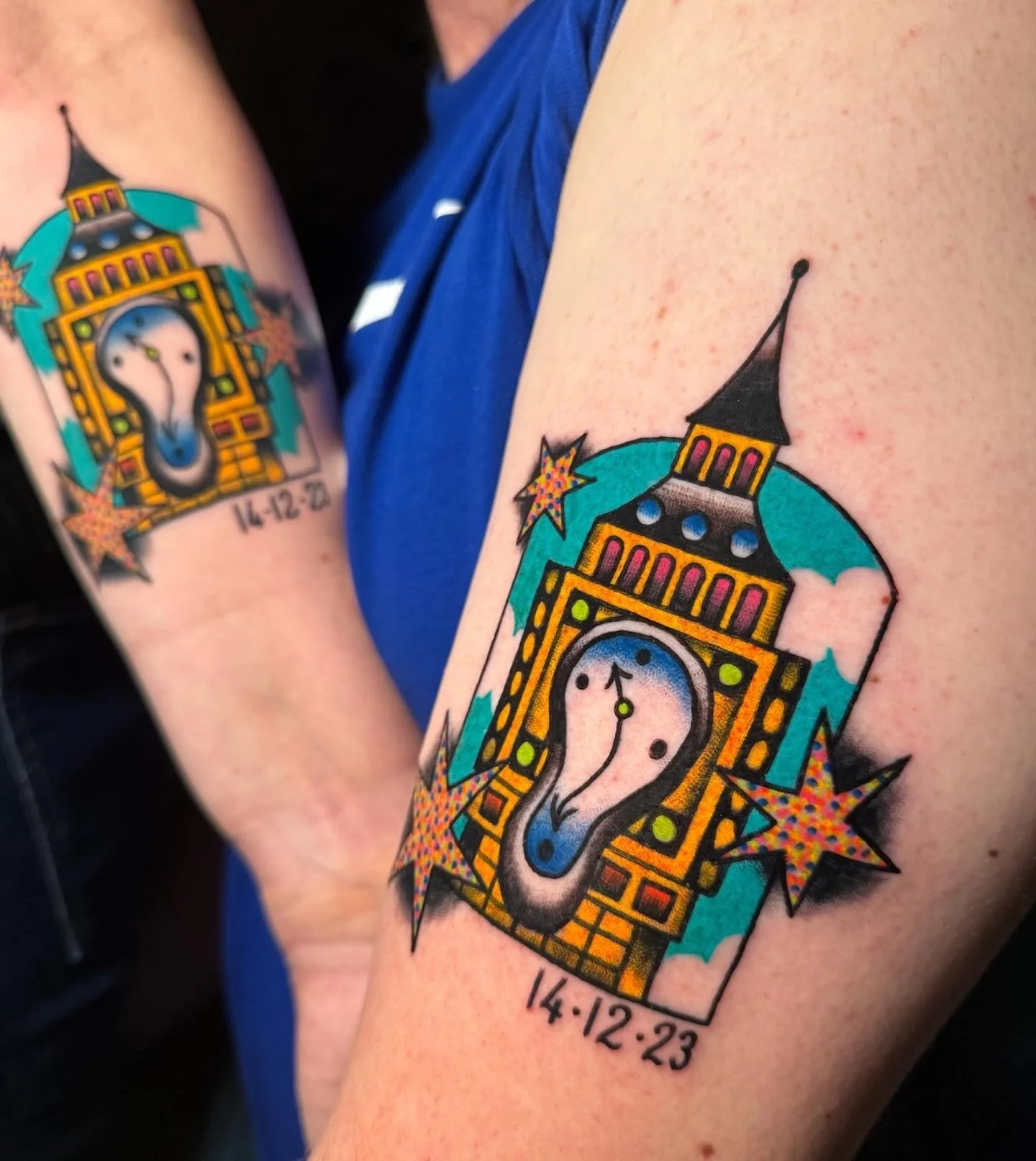 Tattoo of Big Ben clock tower with melting clock face, surrounded by colorful stars, set against a blue background, with the date 14-12-23 below.