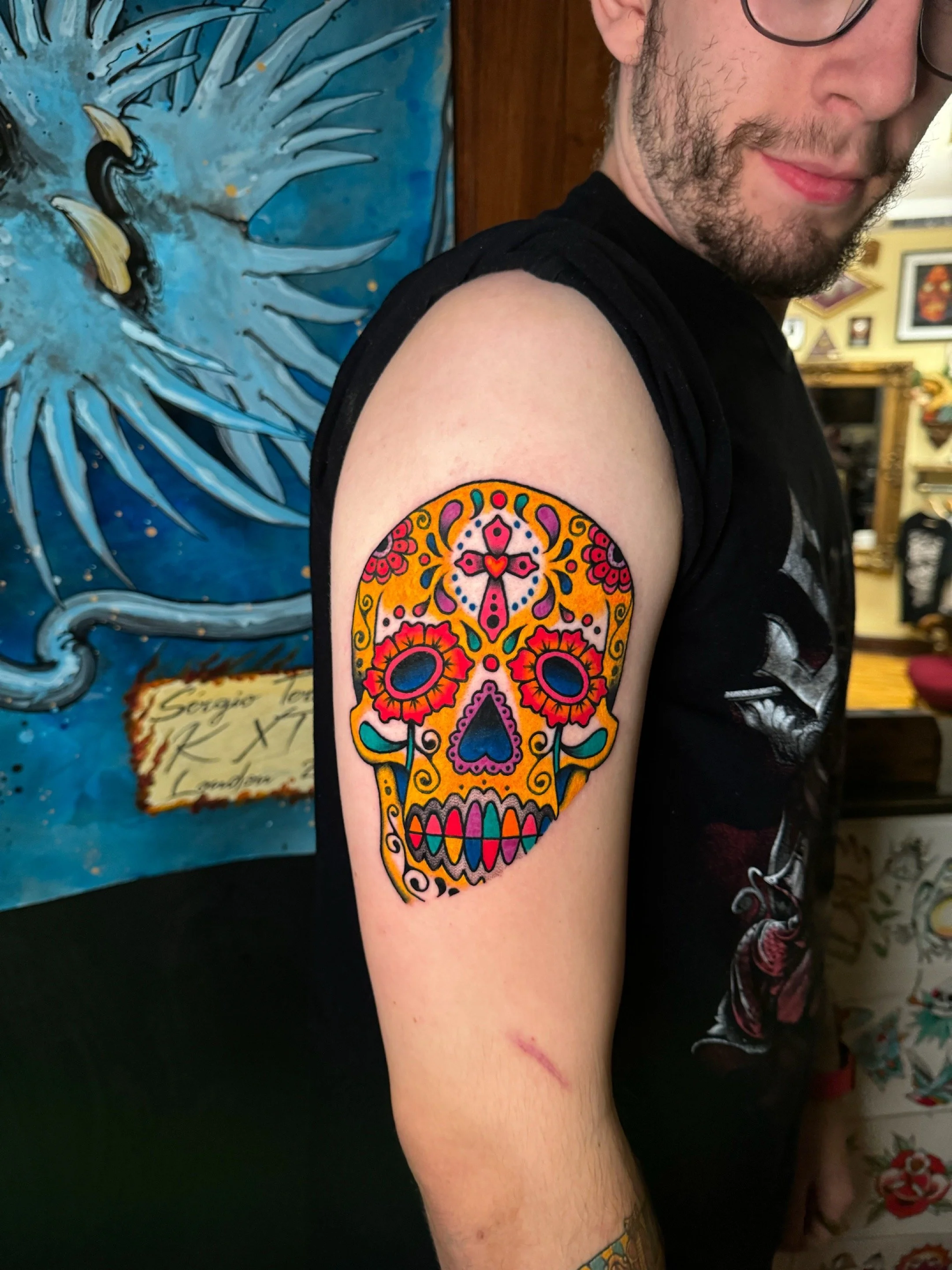 Colorful sugar skull tattoo on person's upper arm, featuring floral and swirl designs in red, yellow, pink, purple, and teal.