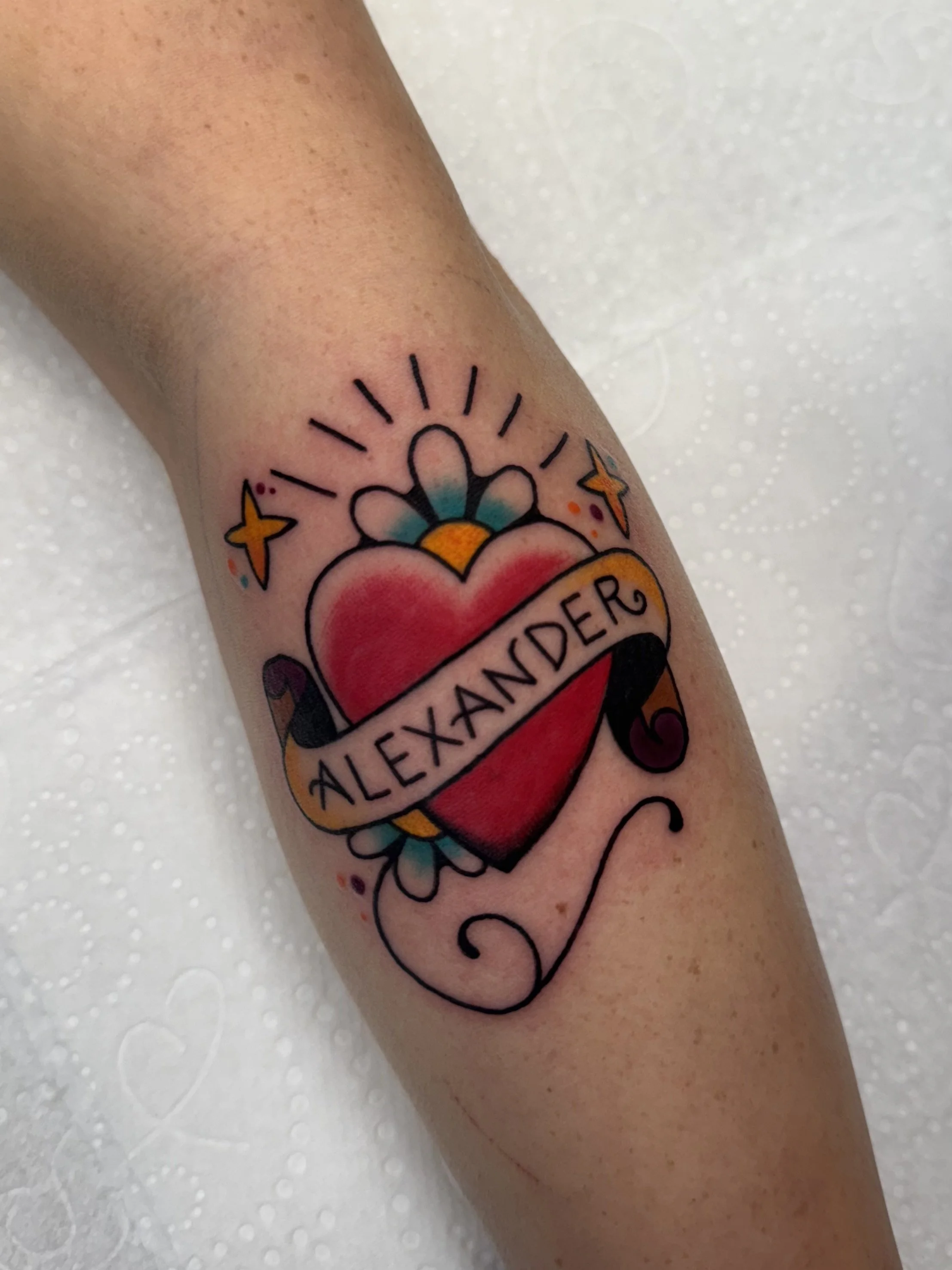 Traditional Tattoo of a red heart with a banner that reads 'ALEXANDER', a blue and yellow flower behind the heart, surrounded by rays, sparkle stars, and decorative swirls.