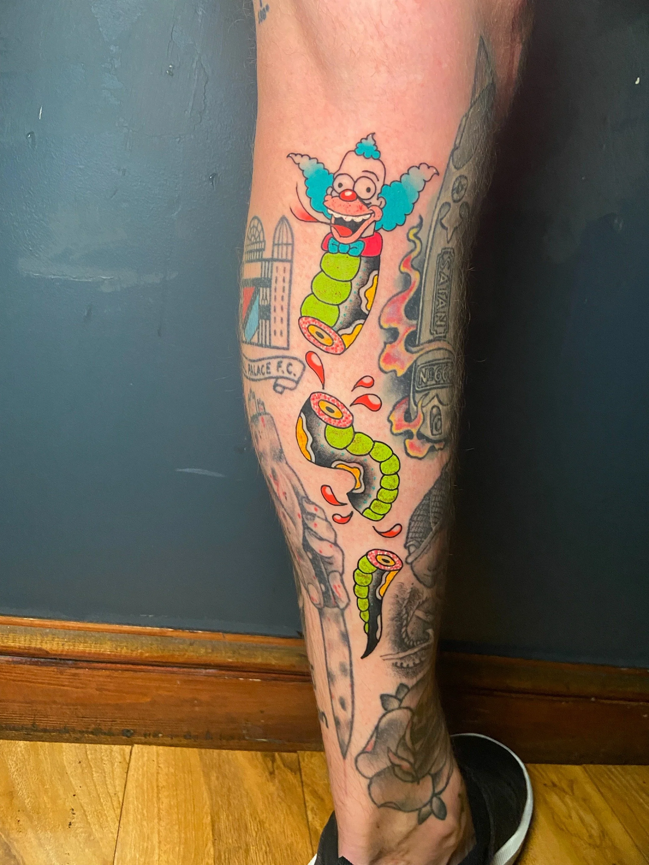 Colorful tattoo of  cartoon clown Krusty the Clown from the Simpsons with blue hair, glasses, and a red nose, merged with a traditional tattoo style snake with a black and green body, on a person's leg.