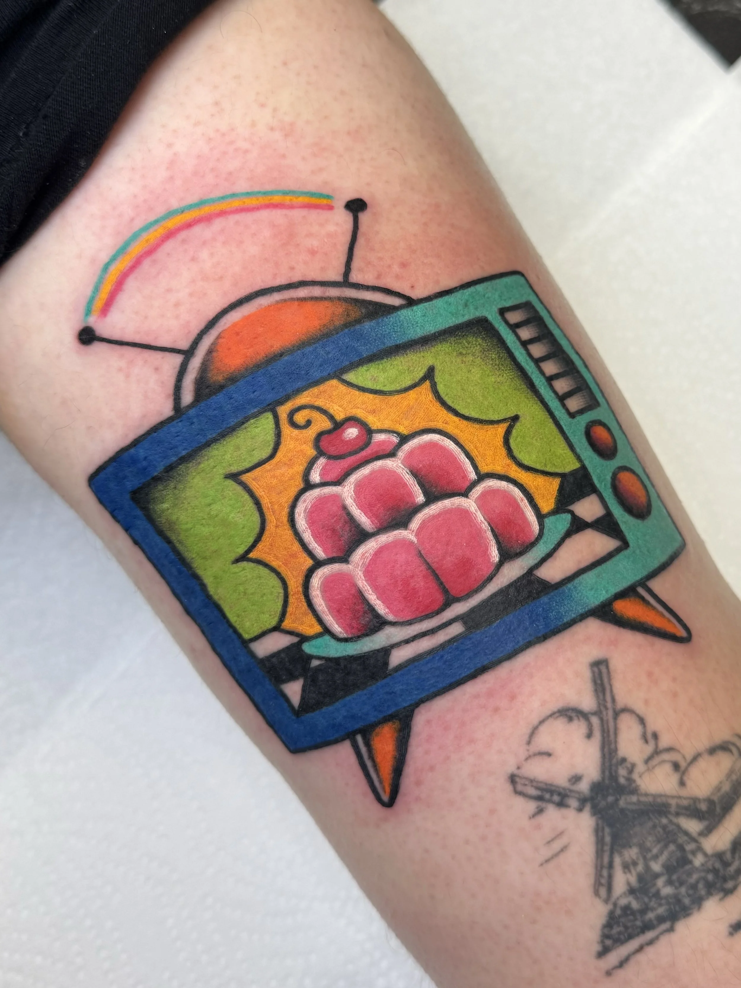 Colorful tattoo of a vintage TV displaying a vintage jello desert with a cherry on top, surrounded by bright orange background, with an antenna on top and a rainbow emanating from the antenna.