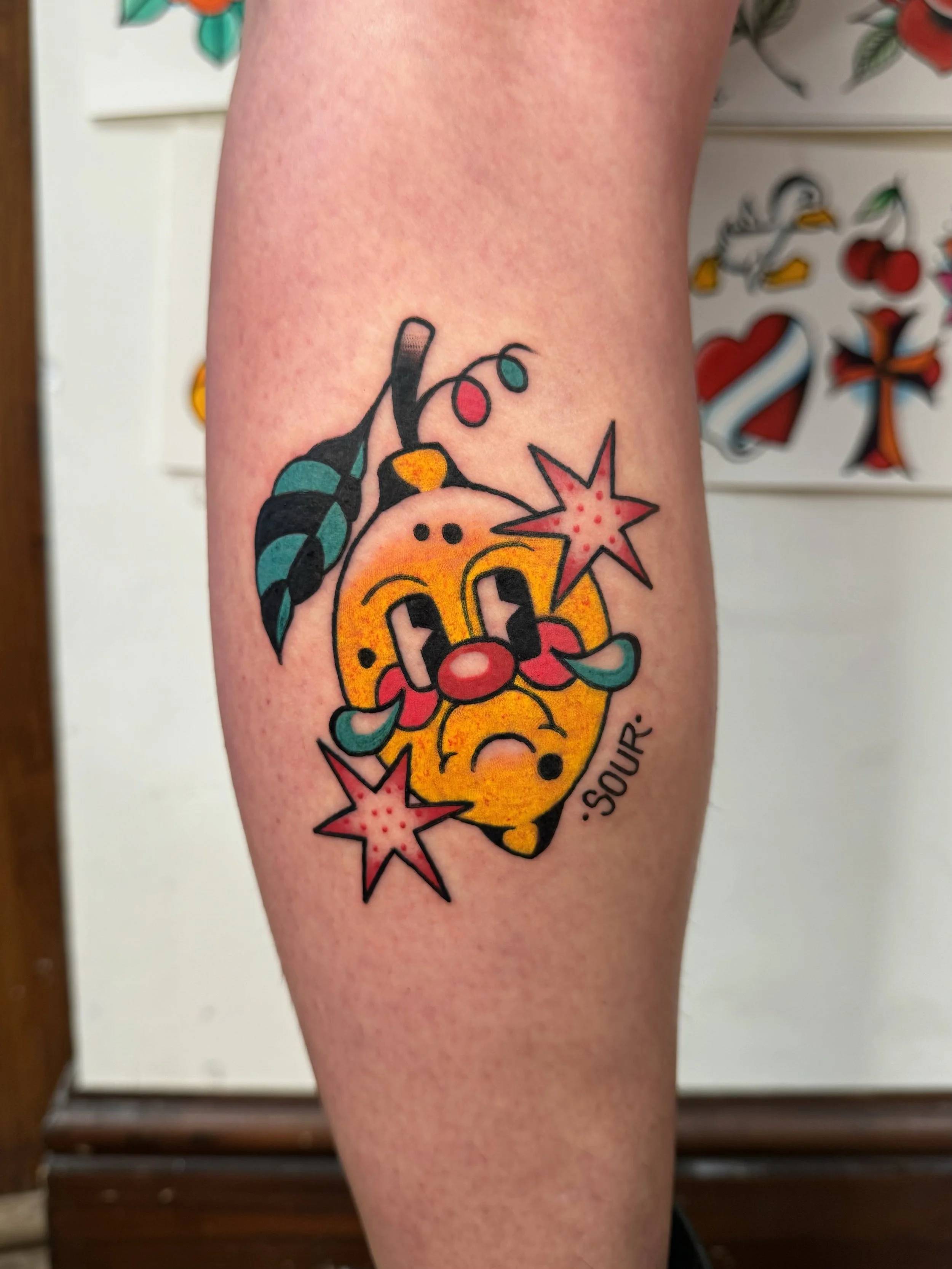 Tattoo of a crying yellow lemon with facial features and a tear, surrounded by cherry blossoms, with the word 'sour' written underneath.