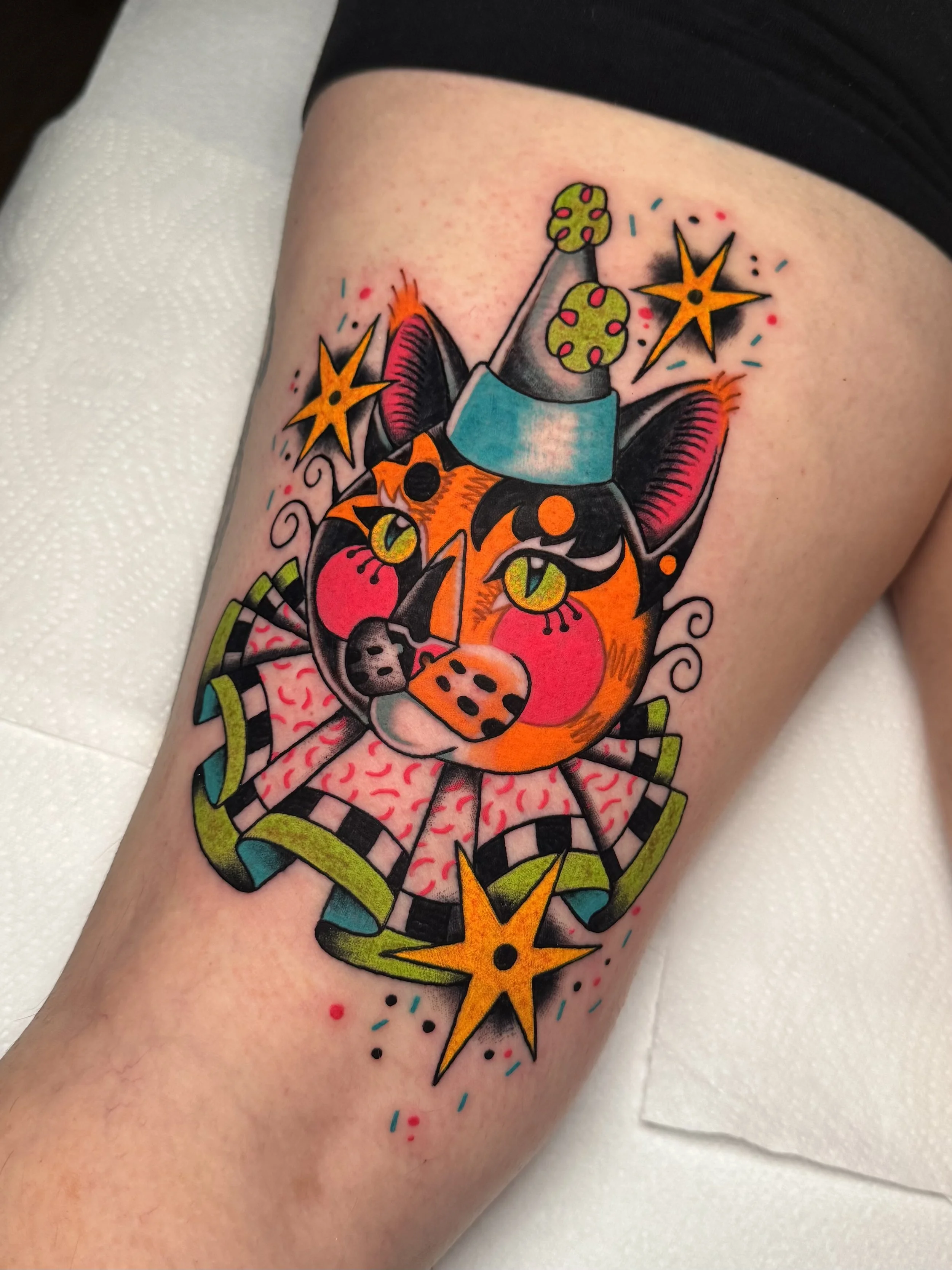 Colorful tattoo of a stylized cat wearing a party hat, with bright orange and black fur, green eyes, pink cheeks, surrounded by stars and confetti-like dots. The cat is a clown.