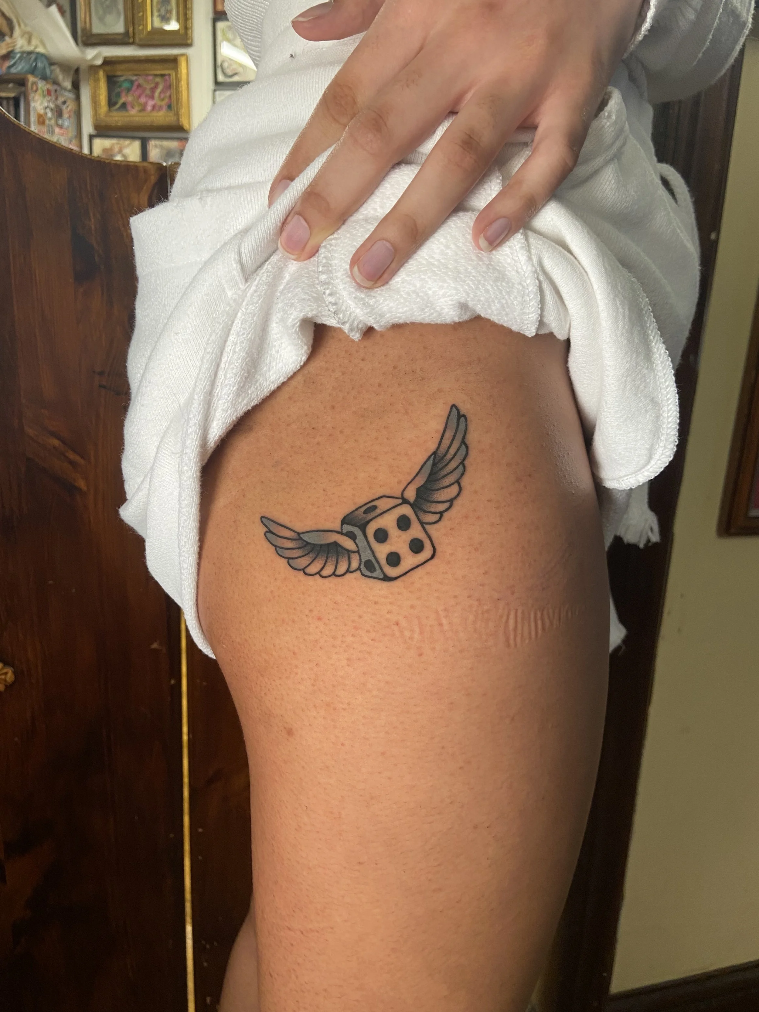 A person's upper thigh reveals a tattoo of a flying dice with wings.