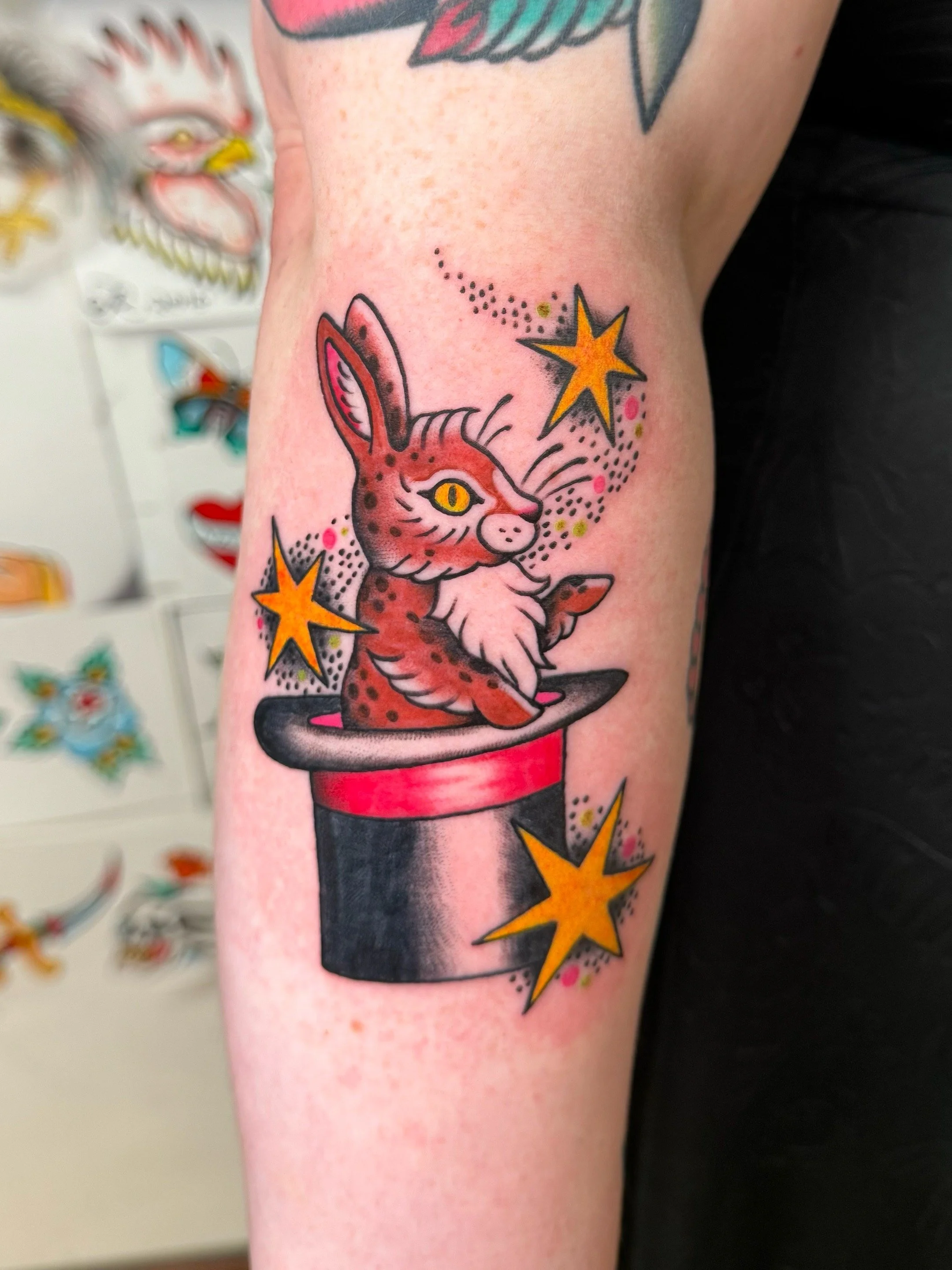 Tattoo of a pink and brown spotted rabbit emerging from a magician's hat, surrounded by yellow stars and colorful dots.