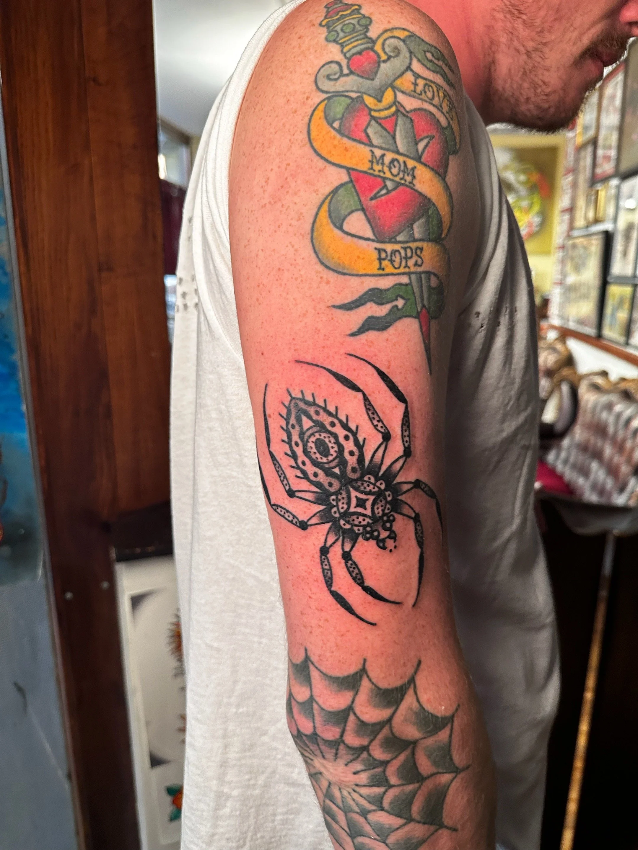Tattoo on man's upper arm depicting a heart with a sword through it, wrapped in a ribbon with the words "LOVE MOM POPS," a spider web, and a spider.