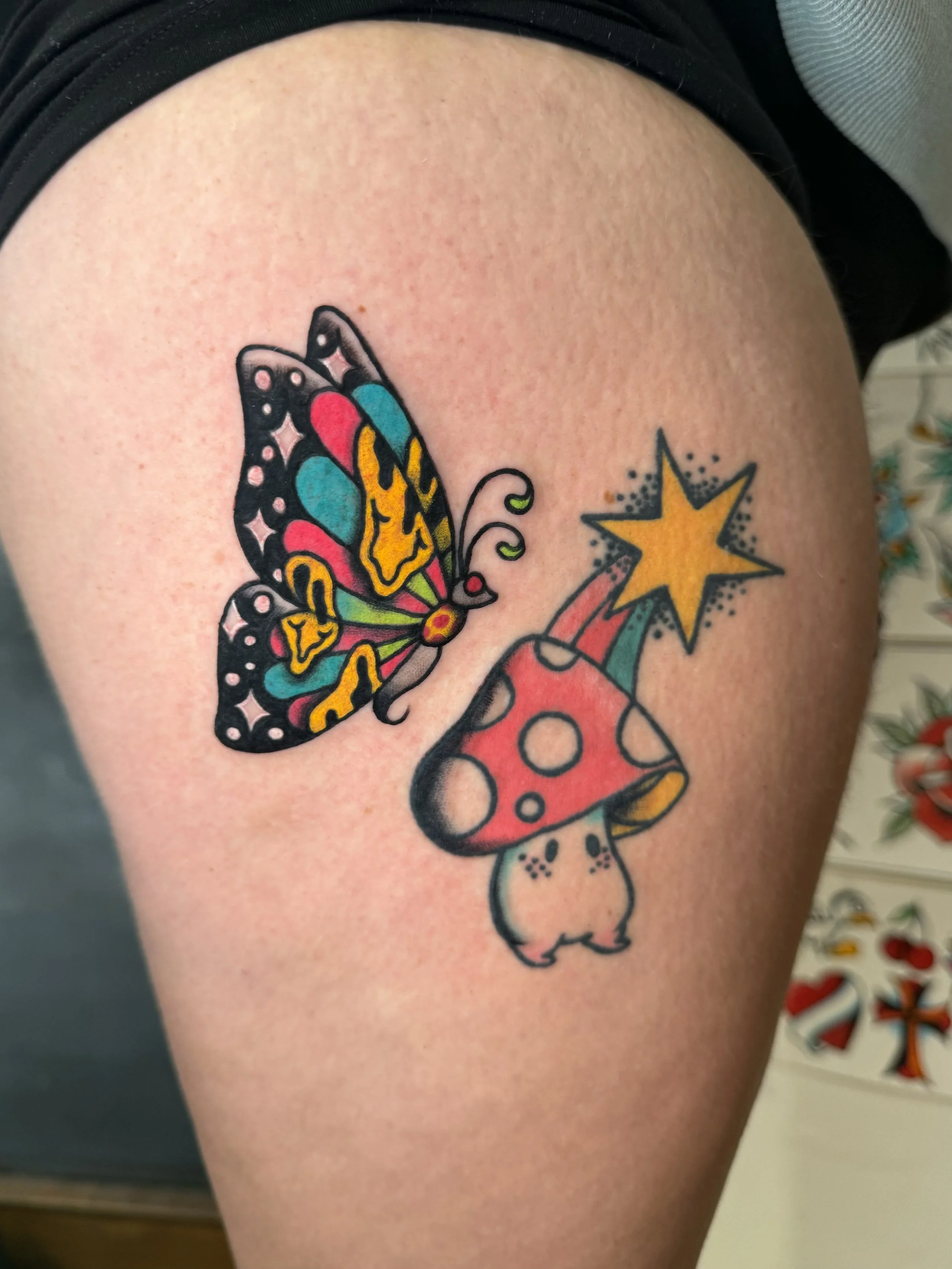 Tattoo of a colorful butterfly, a red and white mushroom, and a yellow star on a person's thigh.