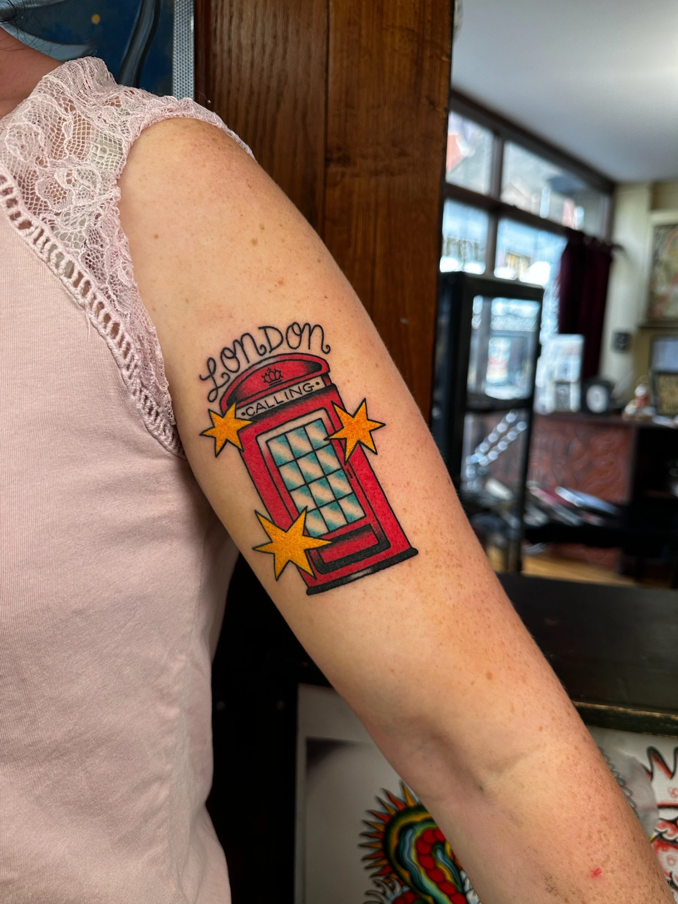 Tattoo of a red telephone booth with the words "London Calling" above, decorated with yellow stars, on a person's upper arm.