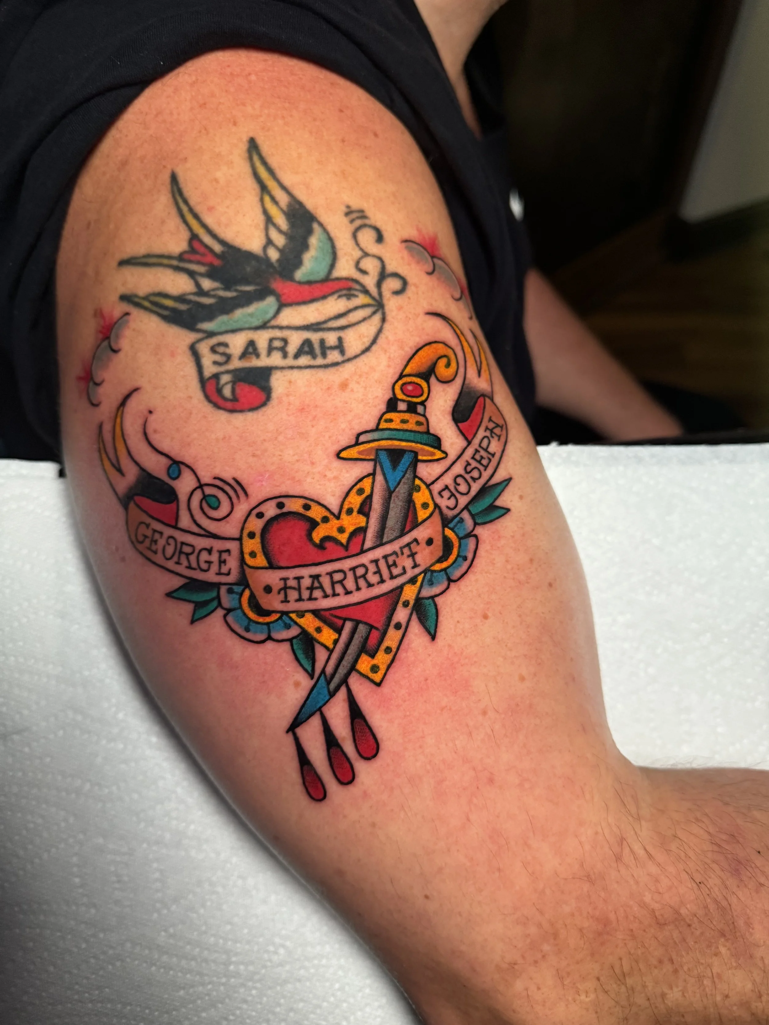 Colorful tattoo on a person's upper arm featuring a swallow bird with a banner that says 'Sarah', a heart with a sword piercing through it, and a ribbon with the names George, Harriet, and Joseph.