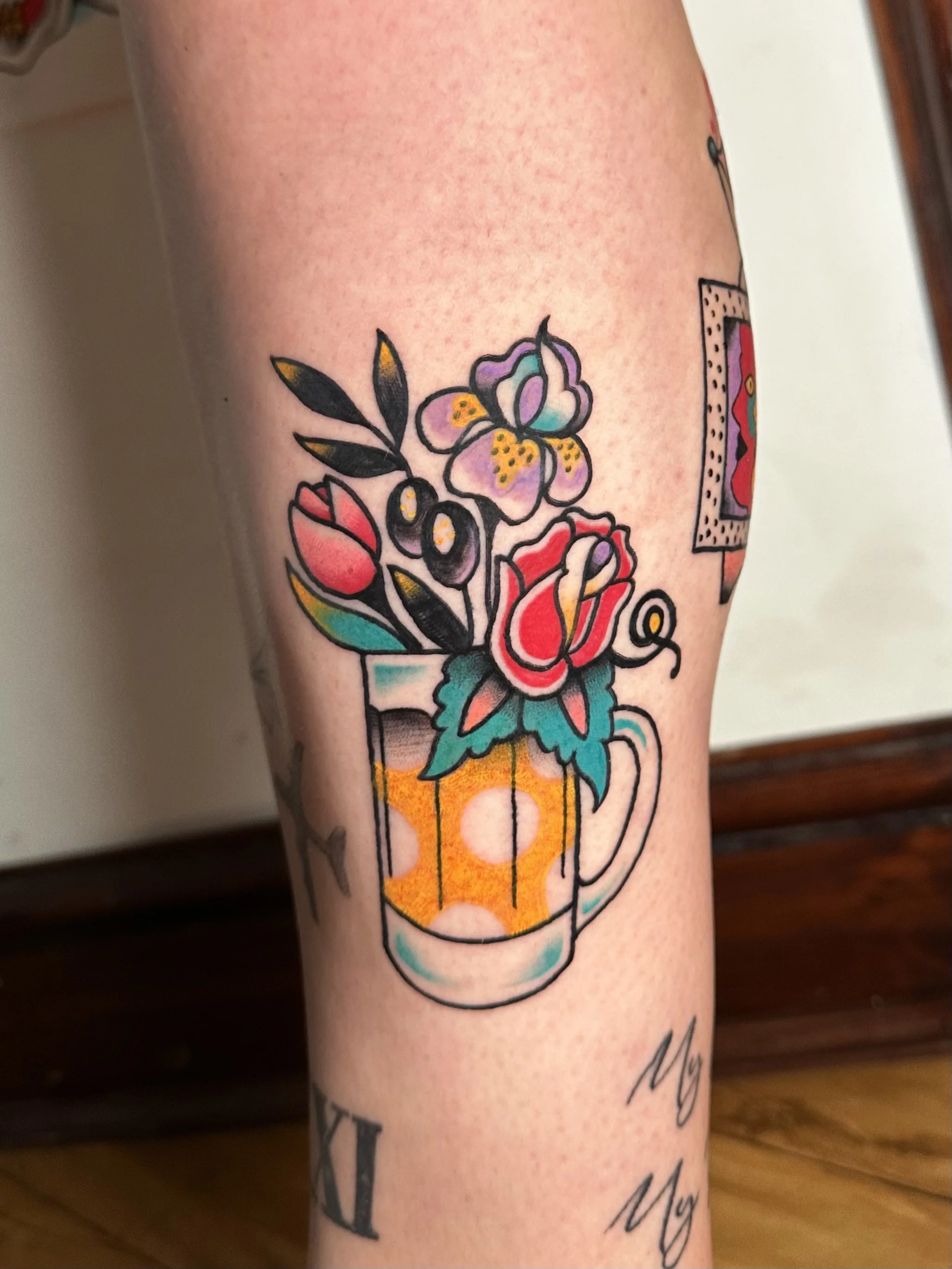 Tattoo of a colorful flower bouquet in a yellow polka-dot pint glass on a person's leg.