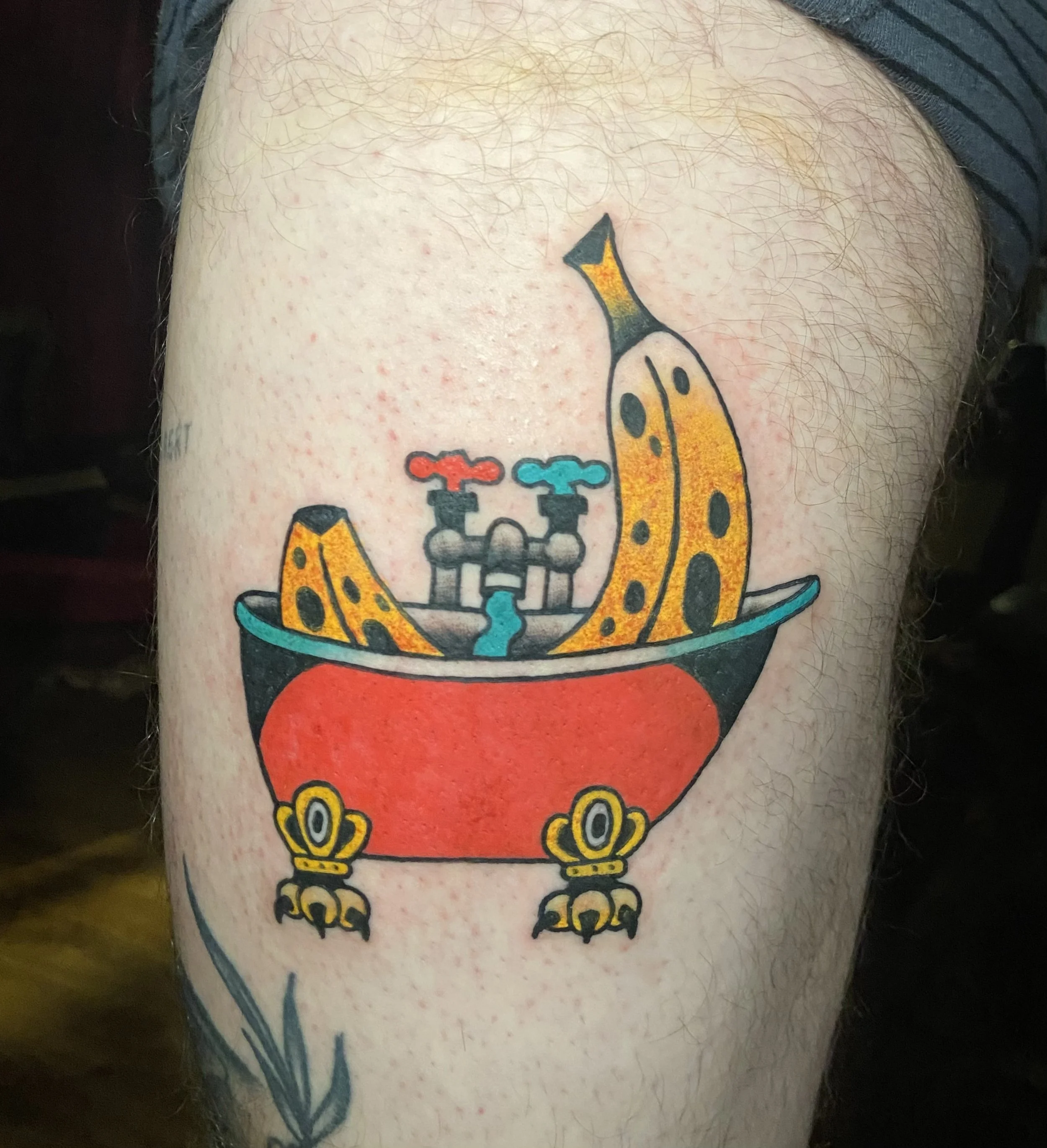 Tattoo of a bathtub with a banana in it, faucets with red and blue handles, on a person's upper thigh.