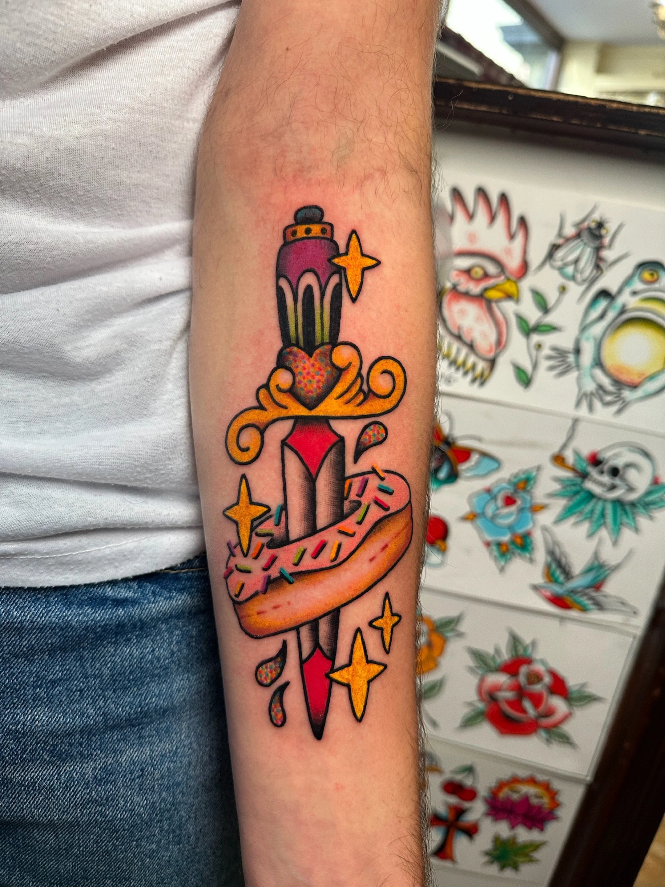 Colorful tattoo of a dagger with a pink and purple handle, a red heart, a donut with colorful sprinkles wrapped around the dagger, surrounded by yellow stars and drops of blood.