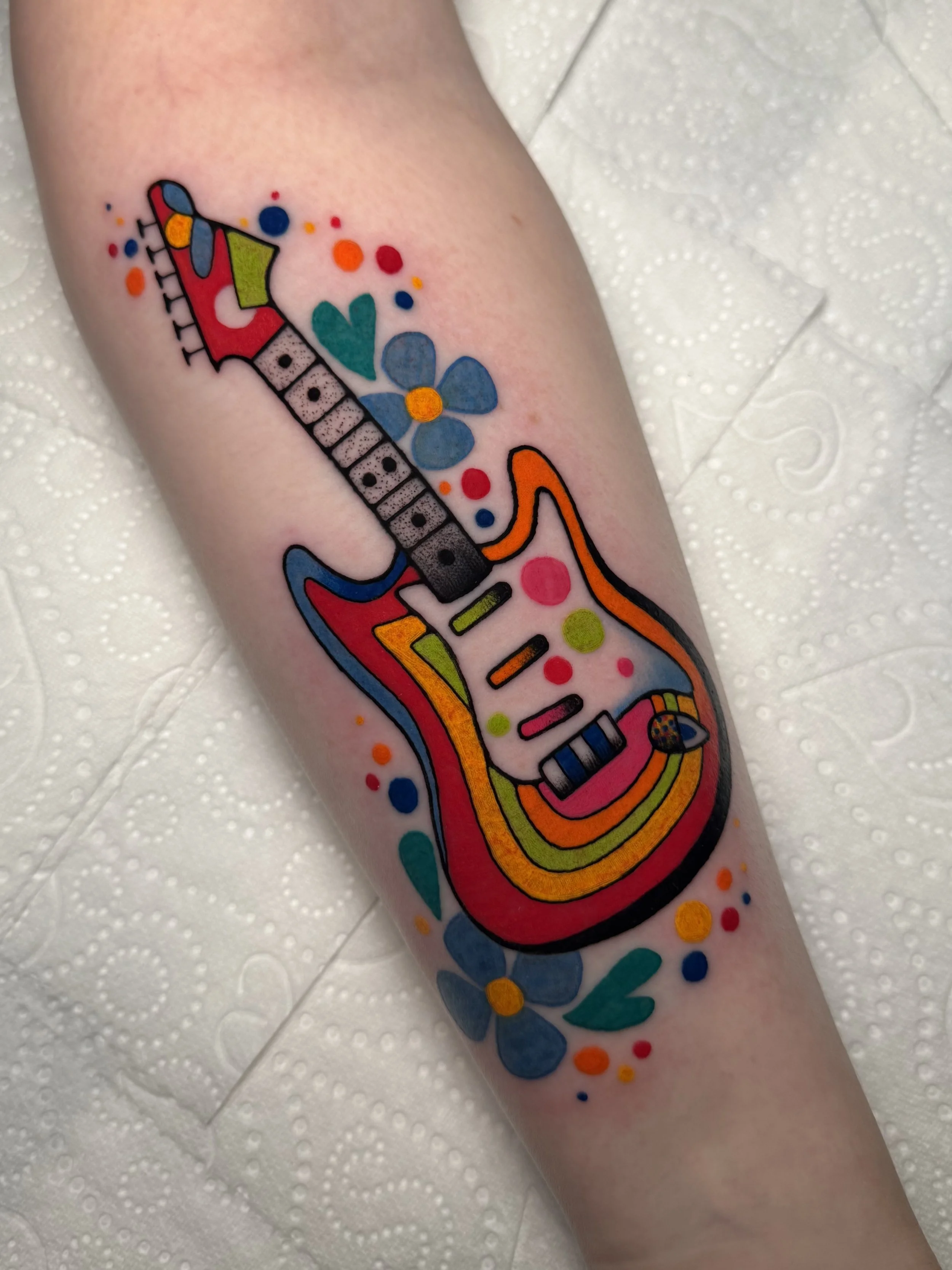 Colorful tattoo of an electric guitar with floral and abstract decorative elements on a person's arm. Guitar is based off of George Harrison on the Beatles' famous rocky guitar.