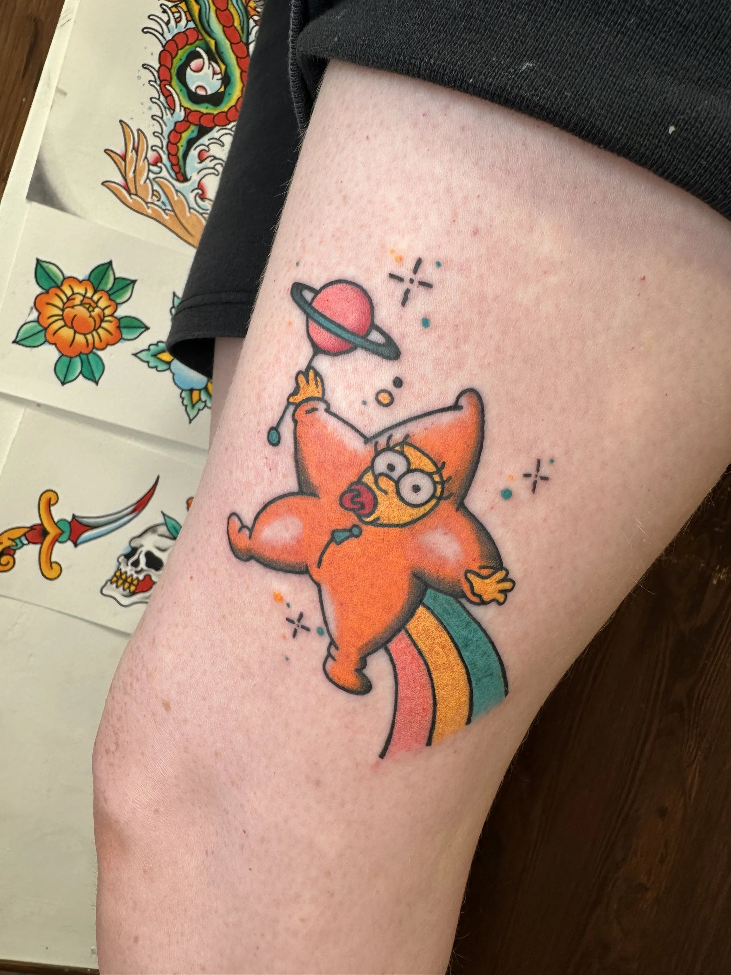 Colorful tattoo of Maggie Simpson in her star suit, holding a planet with rings, standing on a rainbow with stars around.