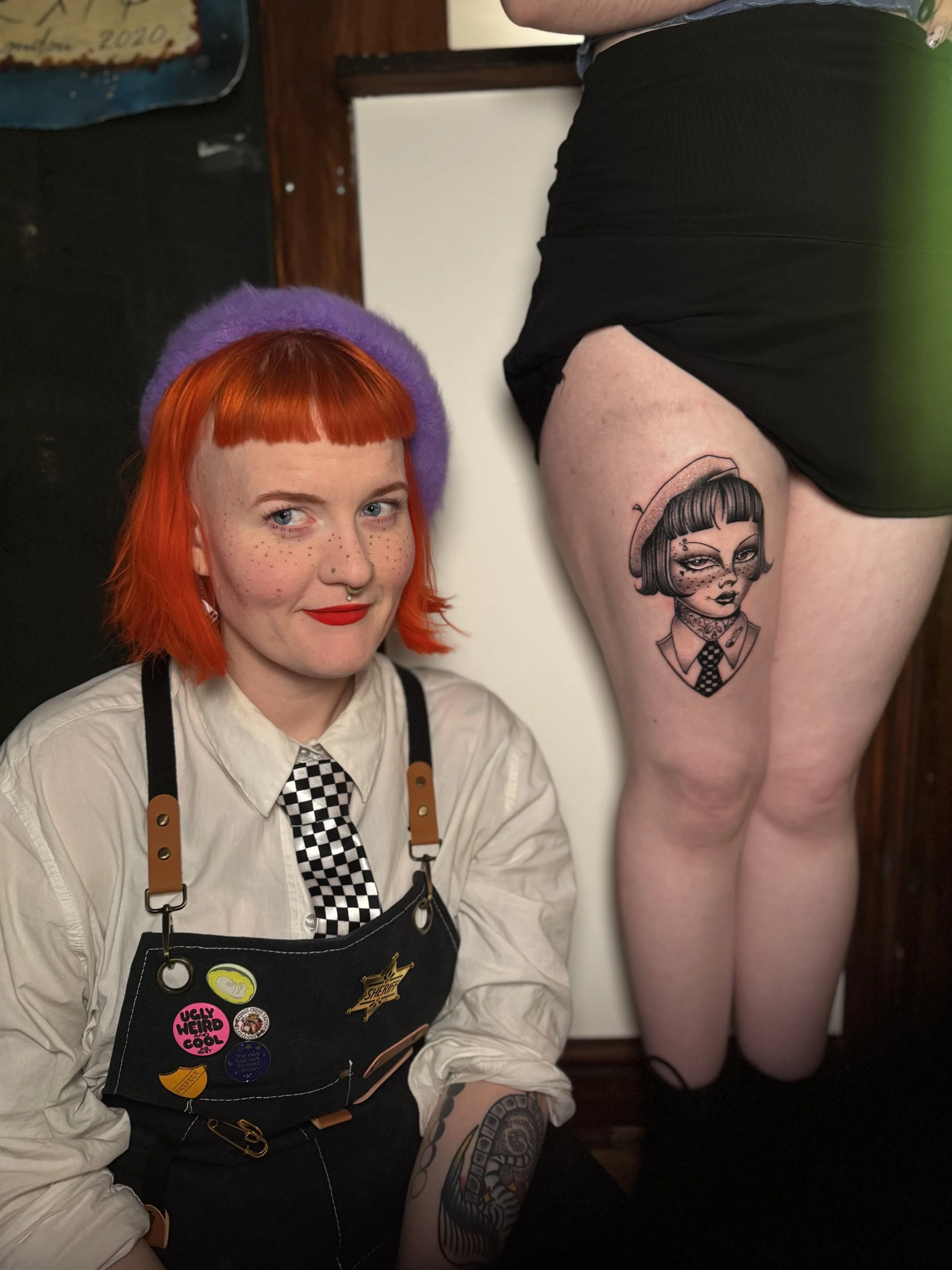 A woman with bright red hair, freckles, and a purple beret sitting indoors next to a person with a tattooed thigh. The woman is wearing a white shirt, checkered tie, and a black apron with buttons and pins. The tattoo depicts a stylized girl with a b