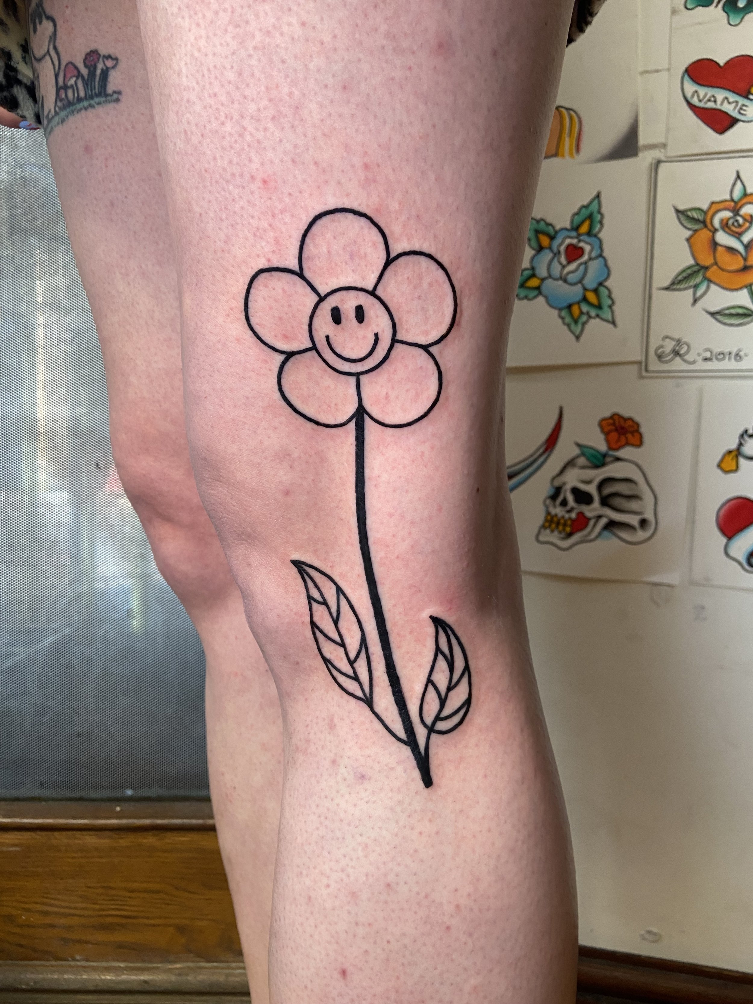 Tattoo of a simple flower with a smiling face, two leaves, and a straight stem on a person's thigh.