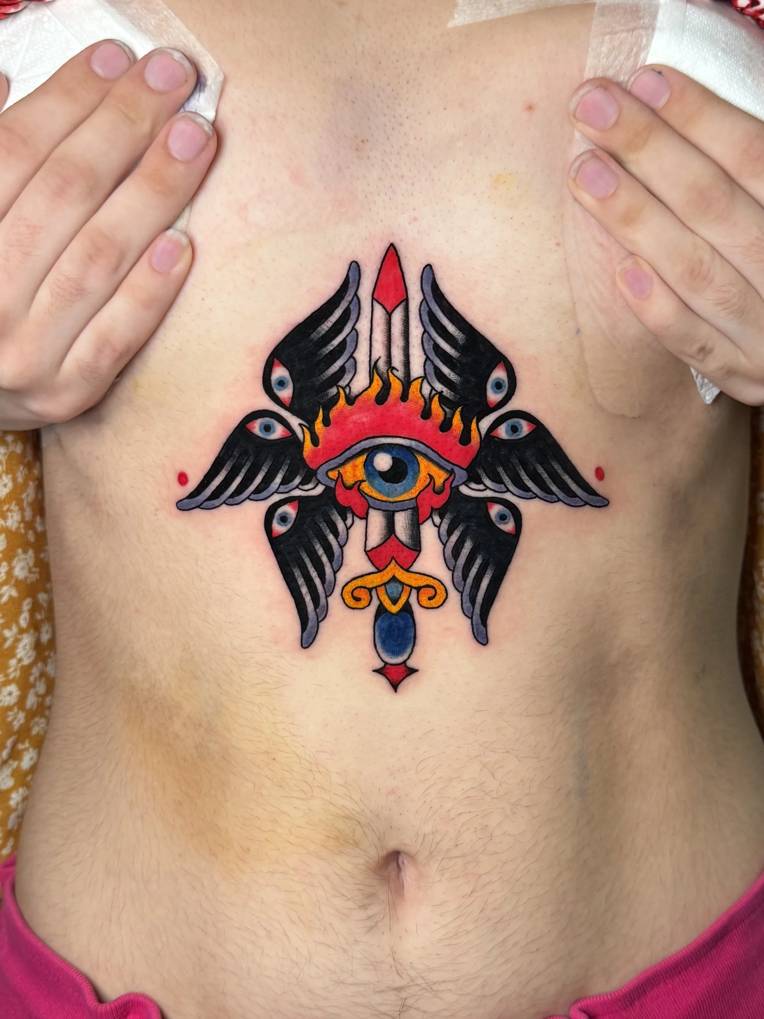 Colorful tattoo of an eye with wings, flames, and a dagger on a person's chest.