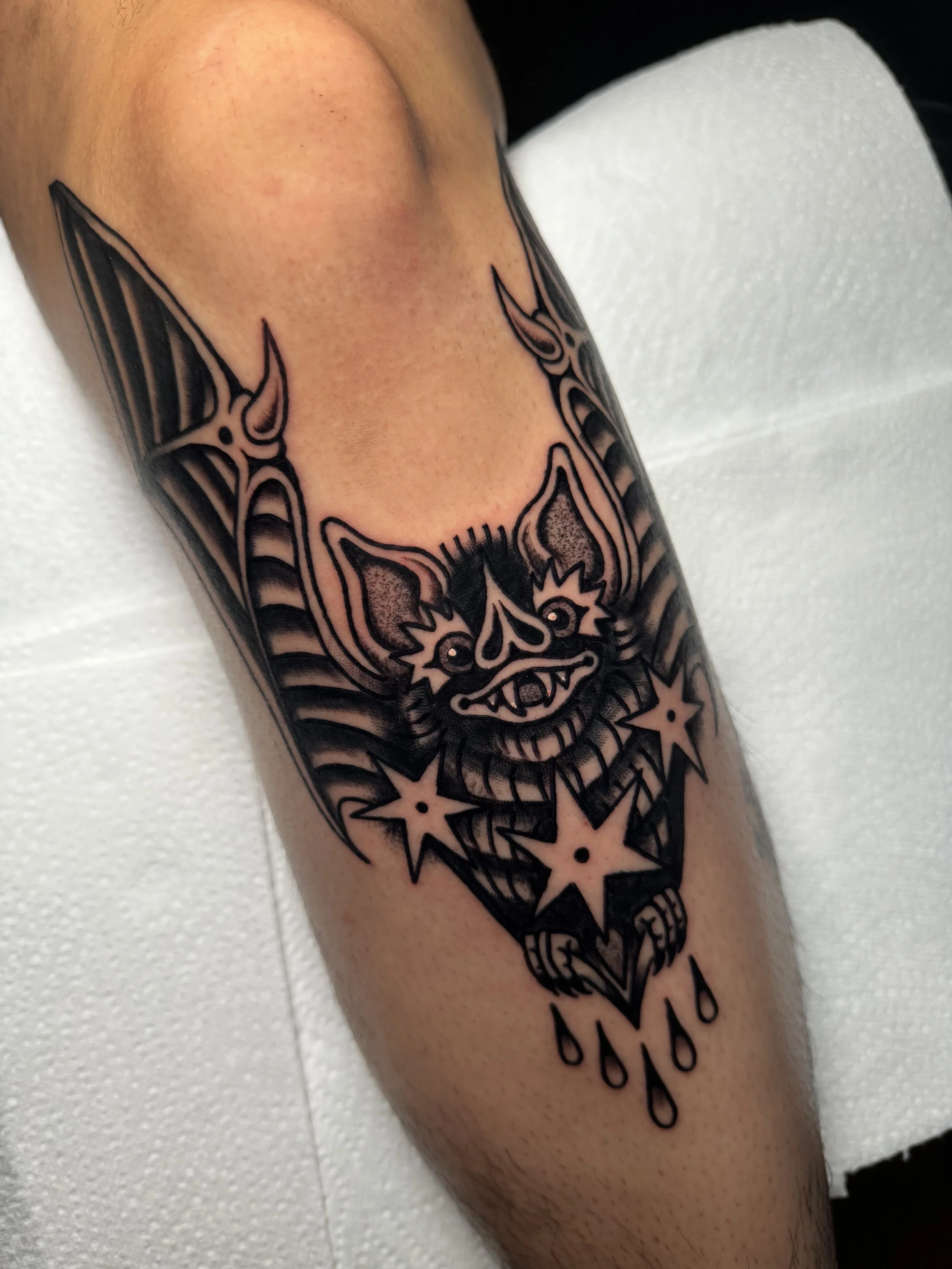 Tattoo of a bat with exaggerated features, large ears, and fanged grin, surrounded by stars and drops, in black ink on a person's knee.