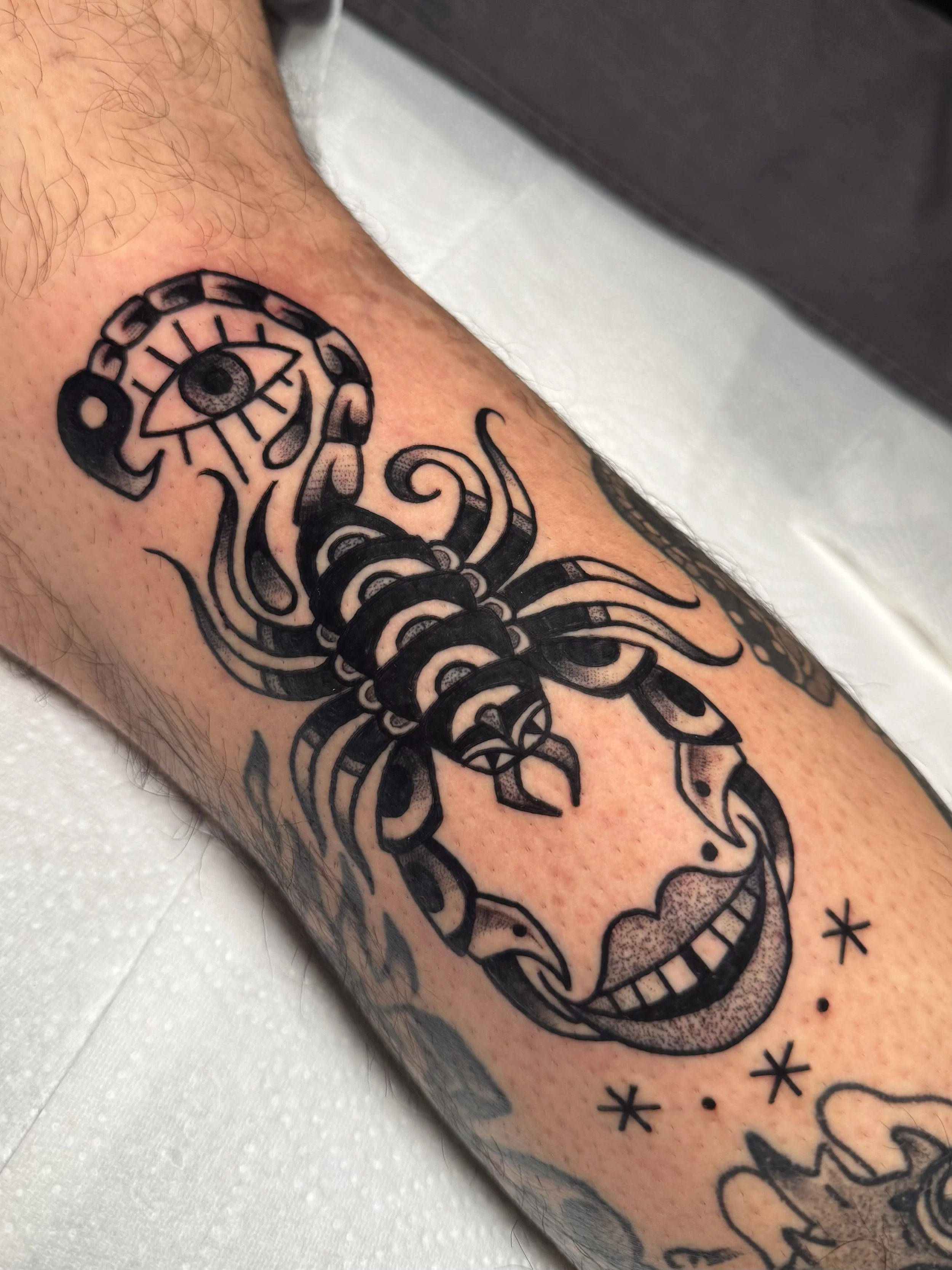 Tattoo of a stylized scorpion with an eye on its tail and a smiling face below, decorated with stars and dots, on a person's arm.