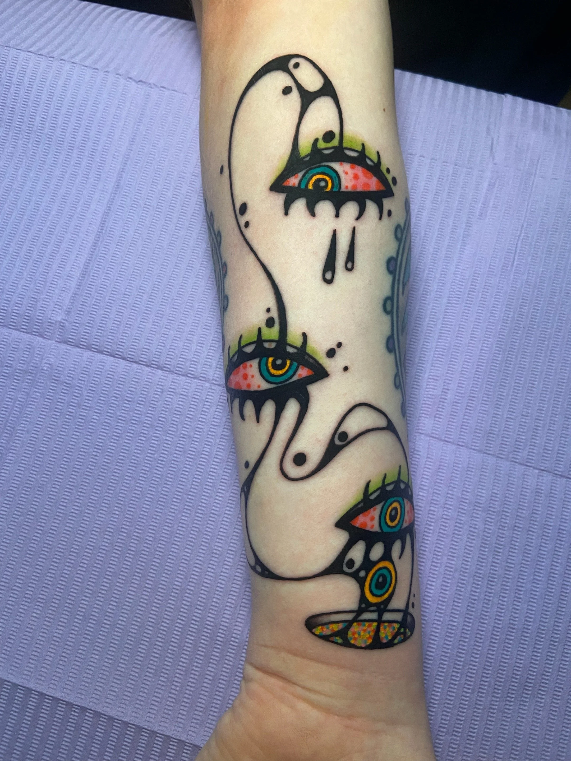 Tattoo of a stylized snake with multiple eyes and a colorful pattern on its head on a person's forearm.