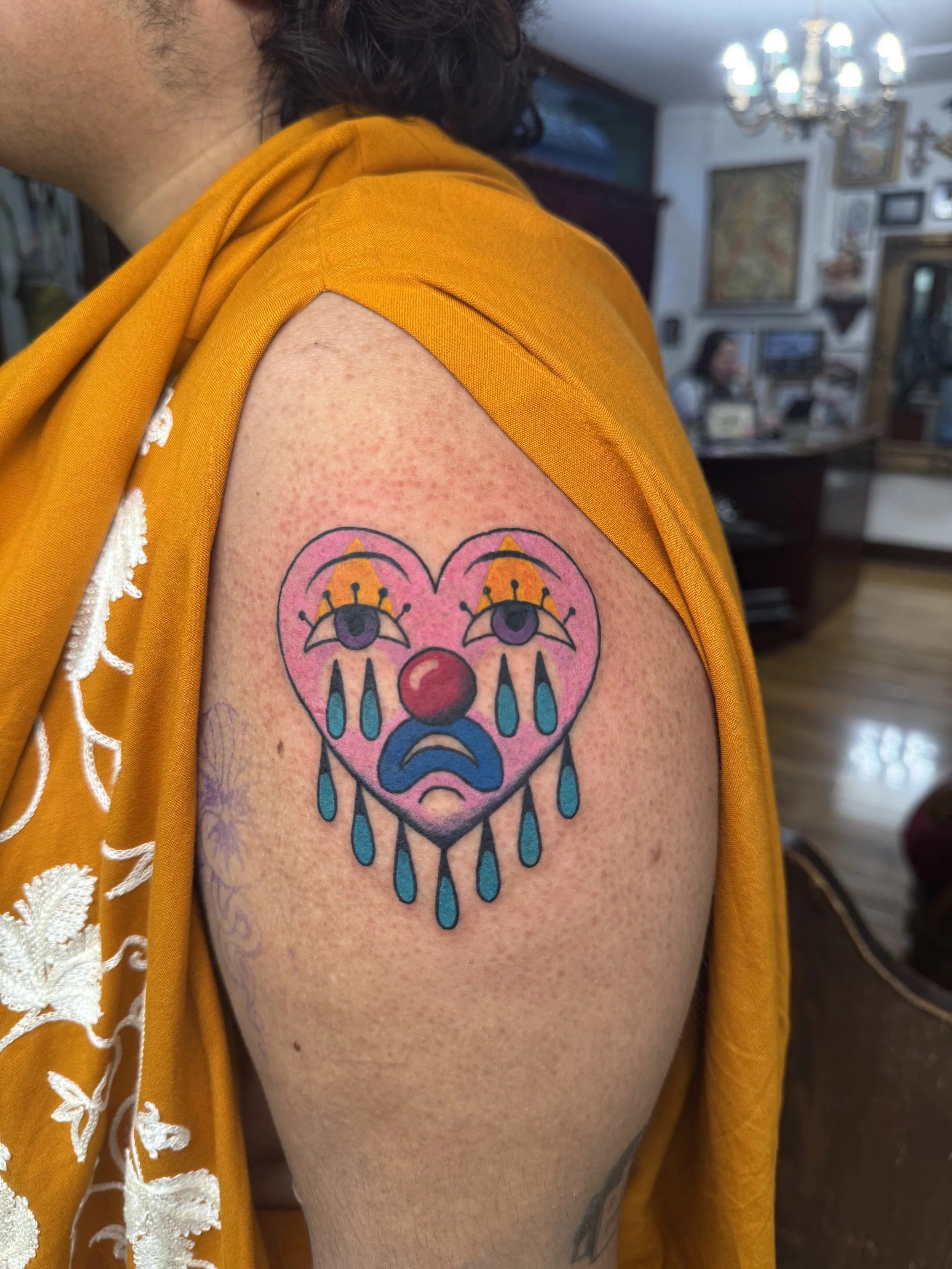 Tattoo of a sad clown face with tears, framed by a heart-shaped outline, on a person's upper arm, with a yellow shirt draped over the shoulder.