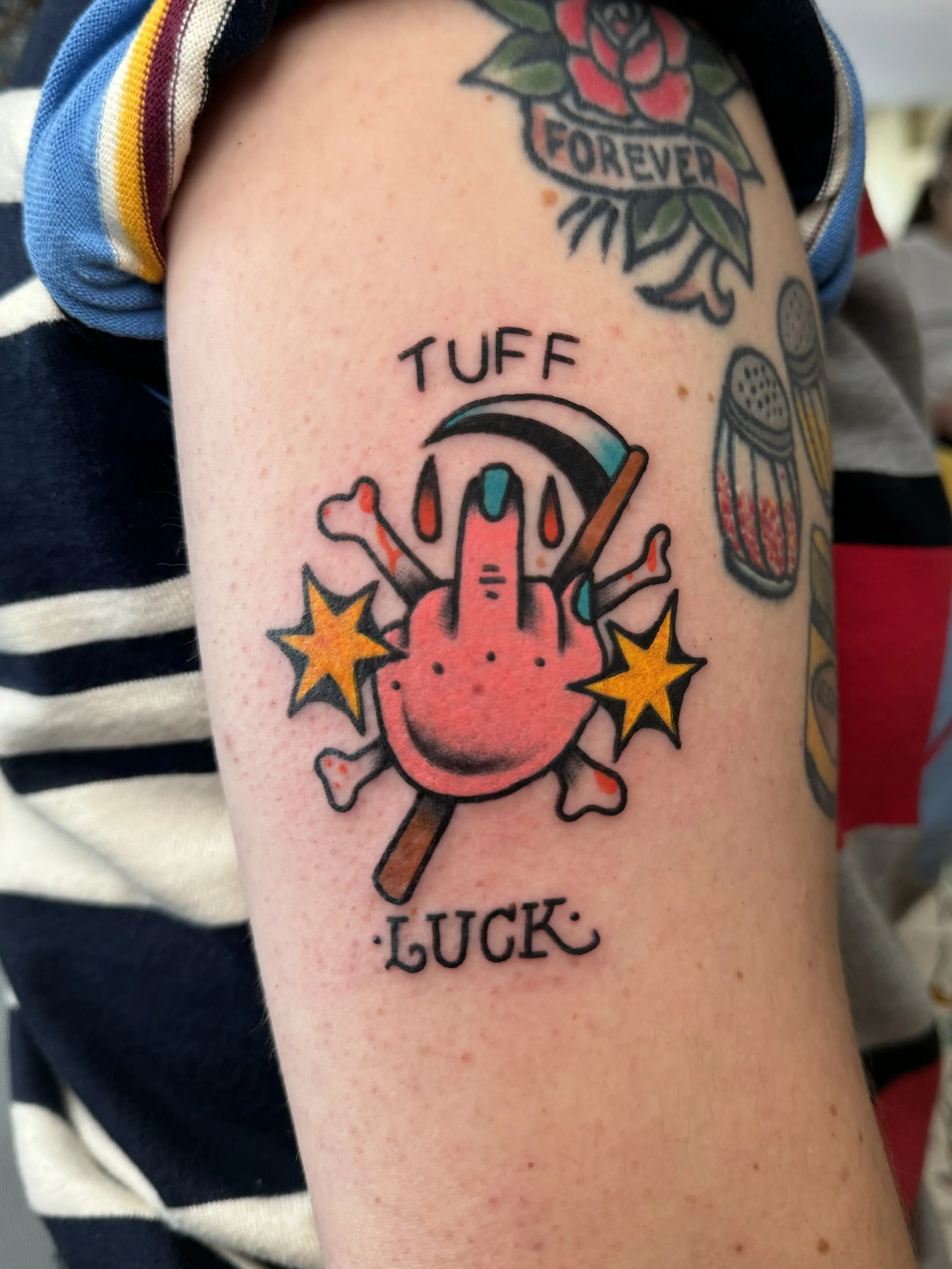 Tattoo of a middle finger  with a scythe and two yellow stars, with the words 'TUFF LUCK' above and below.