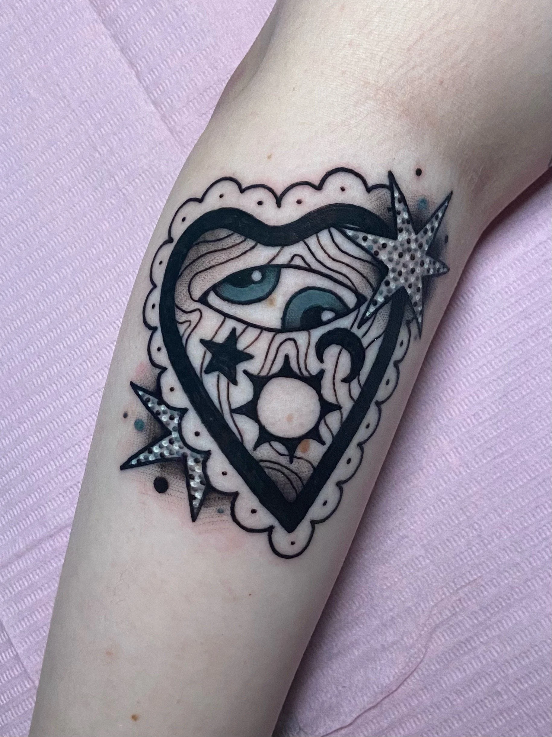 Tattoo of a heart shaped planchette with a wooden texture inside, featuring an eye, stars, crescent moon, and sun symbols, surrounded by a decorative border with star and lightning bolt shapes on a person's arm.