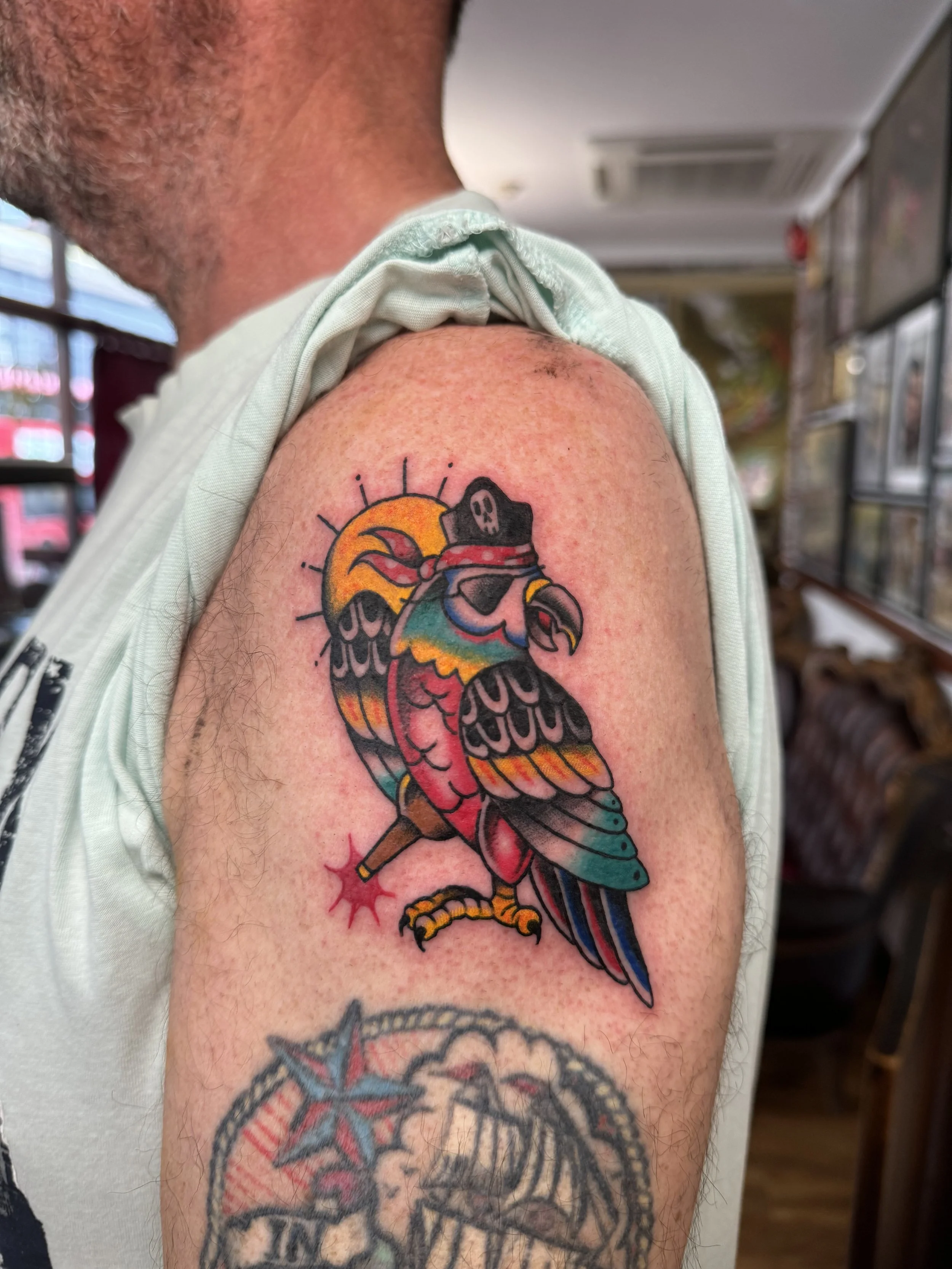 Colorful tattoo of a parrot wearing a pirate hat and sunglasses, with a yellow sun behind it, on a person's upper arm.