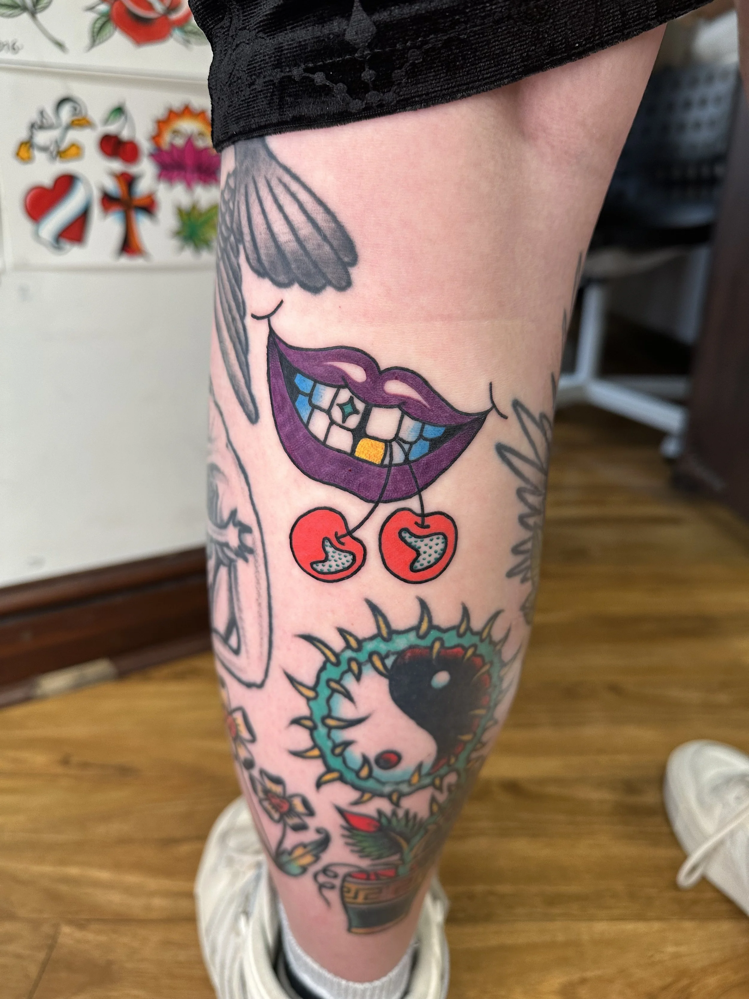 Colorful tattoo on a person's leg featuring a smiling mouth with purple lips and plaid teeth, cherries hanging from the mouth, a yin-yang symbol with spikes, and a flower pot with a blooming plant.